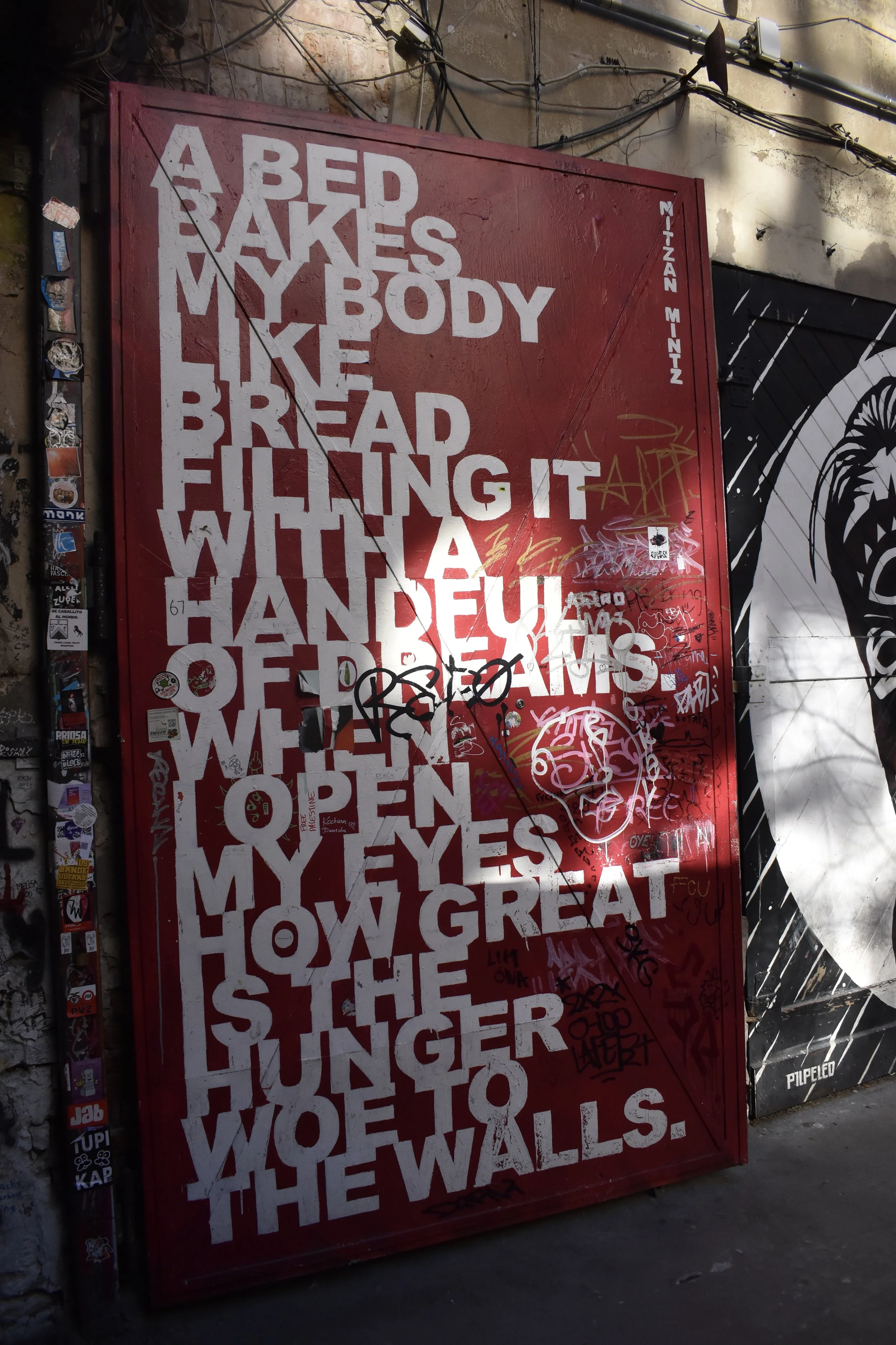 A red poster on a graffiti-covered wall with large white text reading: 'A bed bakes my body like bread, filling it with a handful of dreams. When I open my eyes, how great is the hunger, woe to the walls.' A black and white mural is visible to the ri