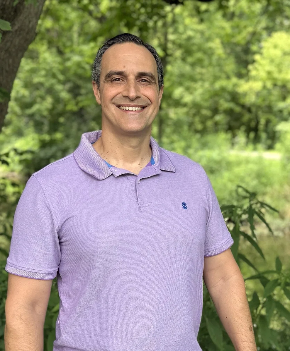 A smiling man in a purple polo shirt standing outdoors with green trees and foliage in the background.