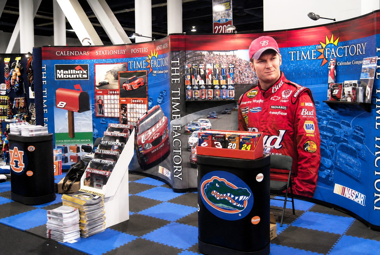 Race car driver in a red racing suit standing in front of a promotional NASCAR display booth with NASCAR memorabilia, posters, and a large graphic of race cars and a driver. The booth features a large background graphic with the logo 'The Time Factory' and an Auburn University logo on a black trash bin.