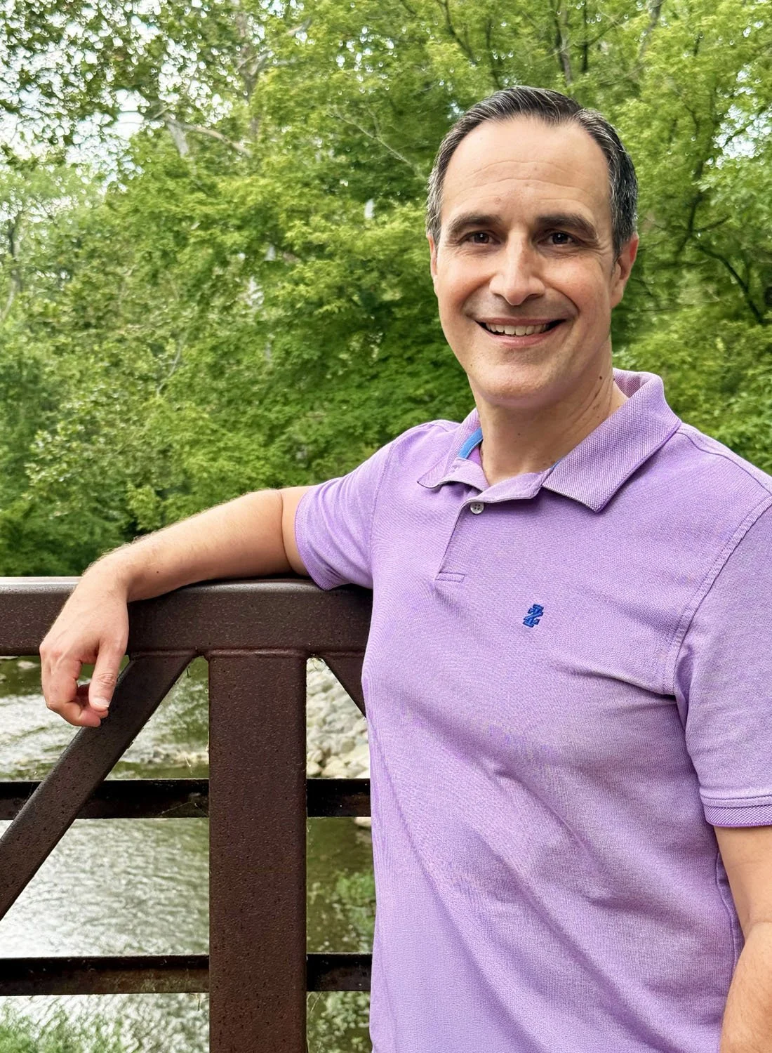A smiling man in a purple polo shirt leaning on a wooden bridge railing outdoors, with green trees and a river in the background.