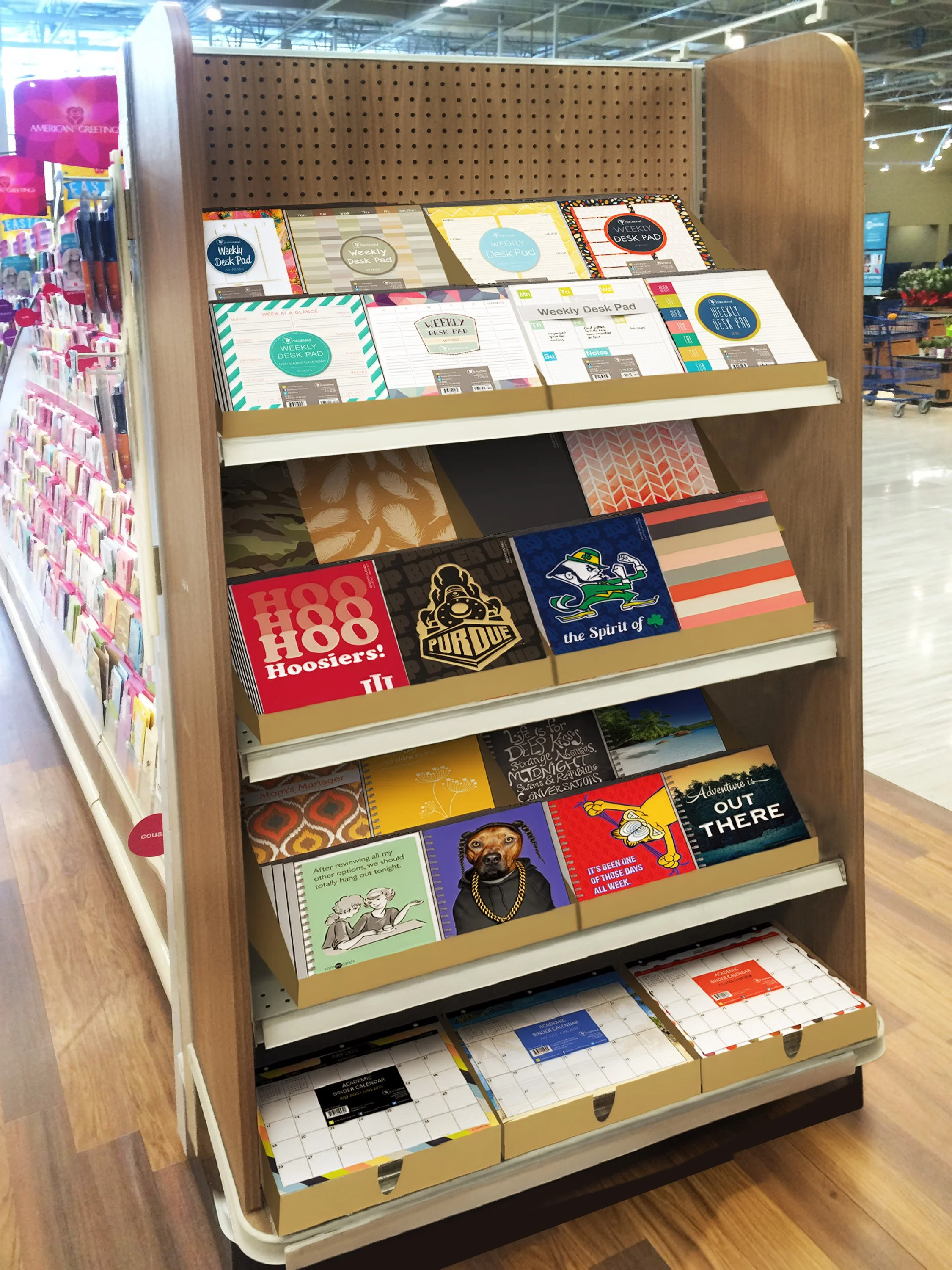 A display shelf in a store with various stationery products, including notebooks, planner pads, and greeting cards. To the left of the shelf are colorful packs of pens or markers. The background shows a store interior with shopping carts and bright lighting.