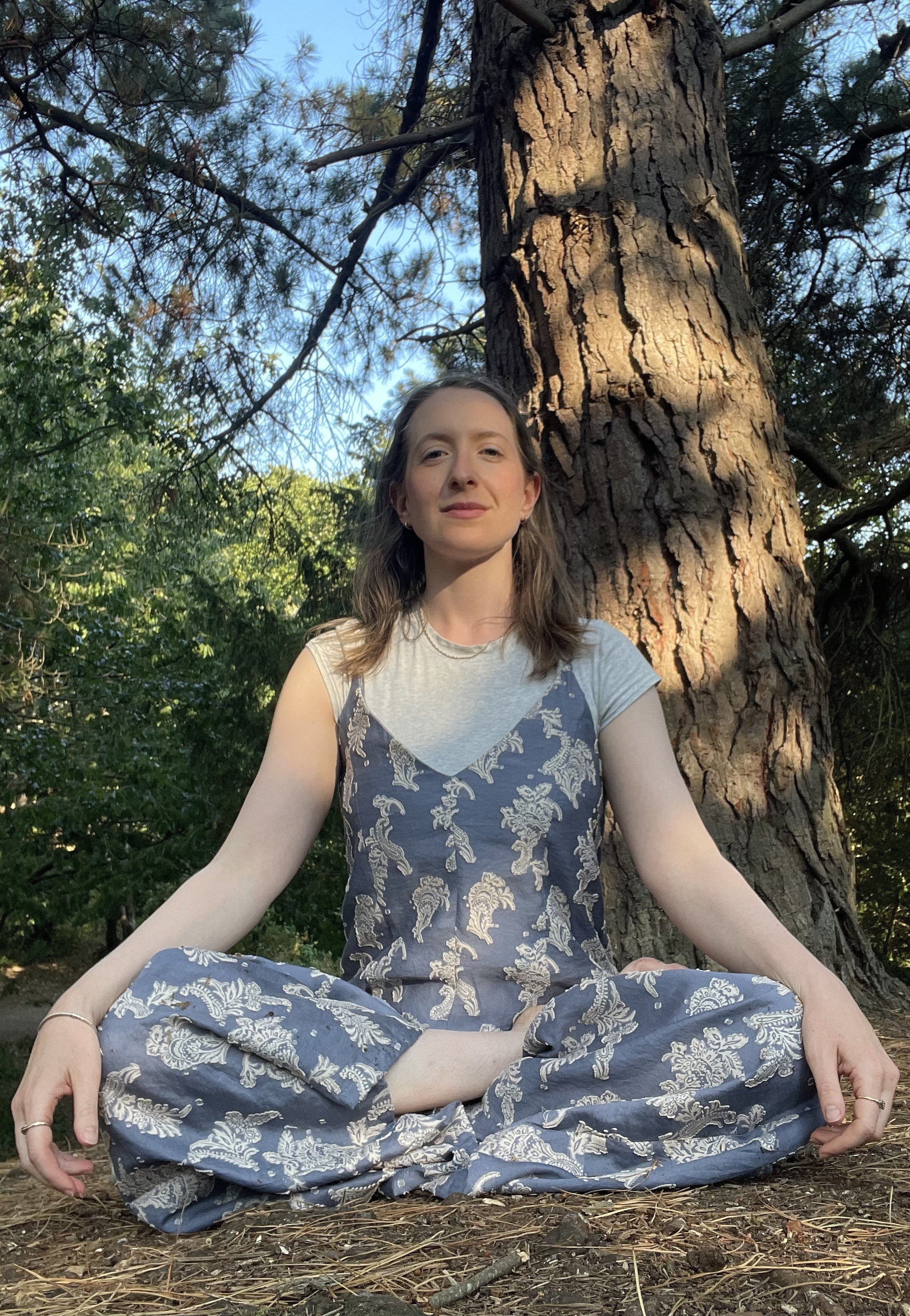 A young woman with brown hair sitting cross-legged on the ground outdoors, meditating near a large tree, wearing a blue patterned dress, with greenery and blue sky in the background.