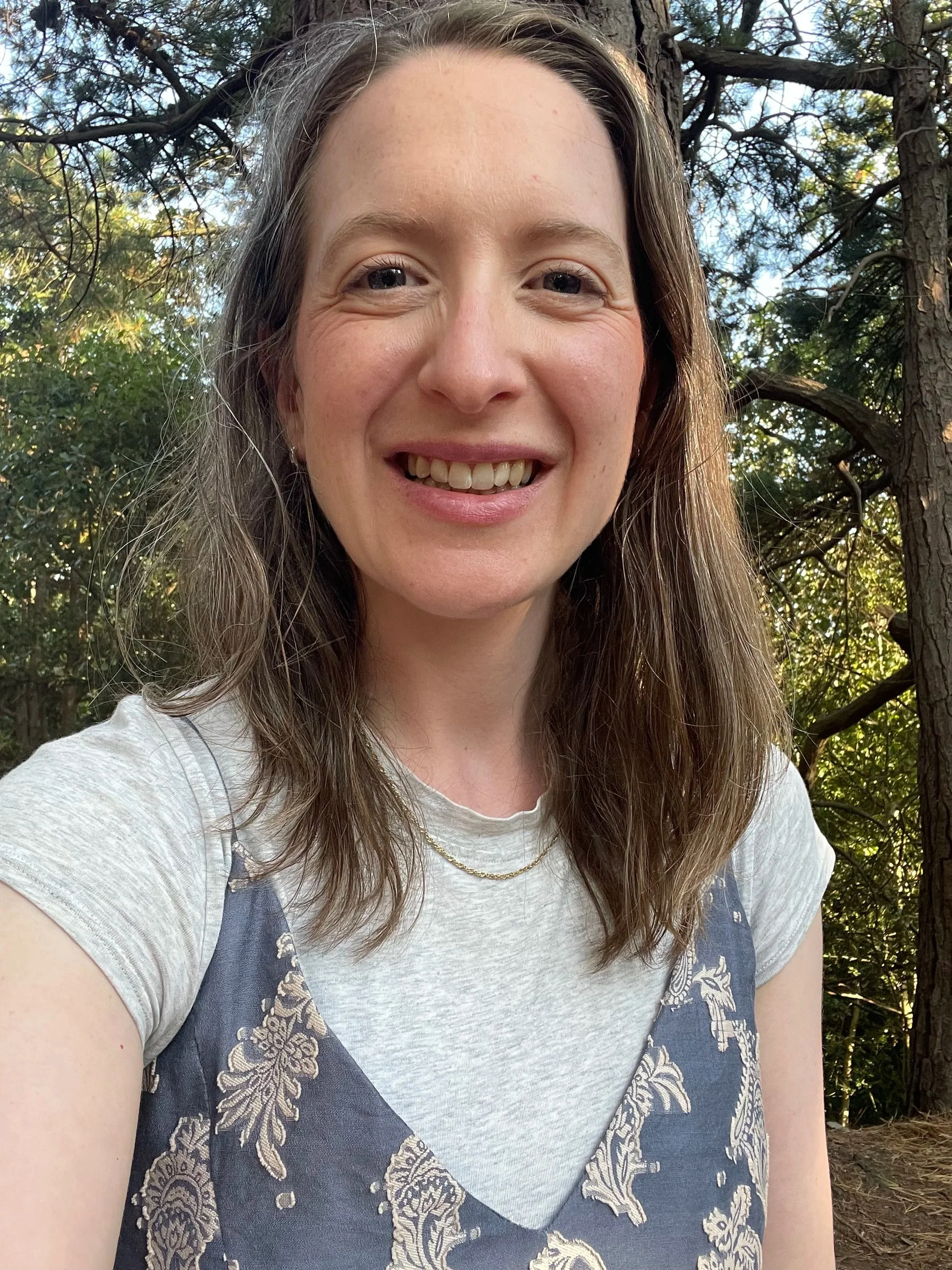A woman with long wavy hair smiling outdoors in a wooded area with sunlight filtering through trees.