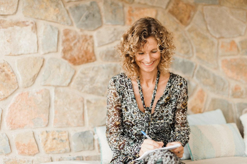 A woman with curly blonde hair, smiling, sitting on a couch with a stone wall in the background, holding a pen and a notepad.