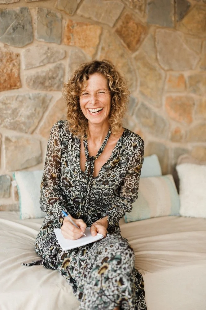 A woman with curly blonde hair, wearing a patterned dress, smiling and holding a notepad and pen while sitting on a bed with stone wall background.