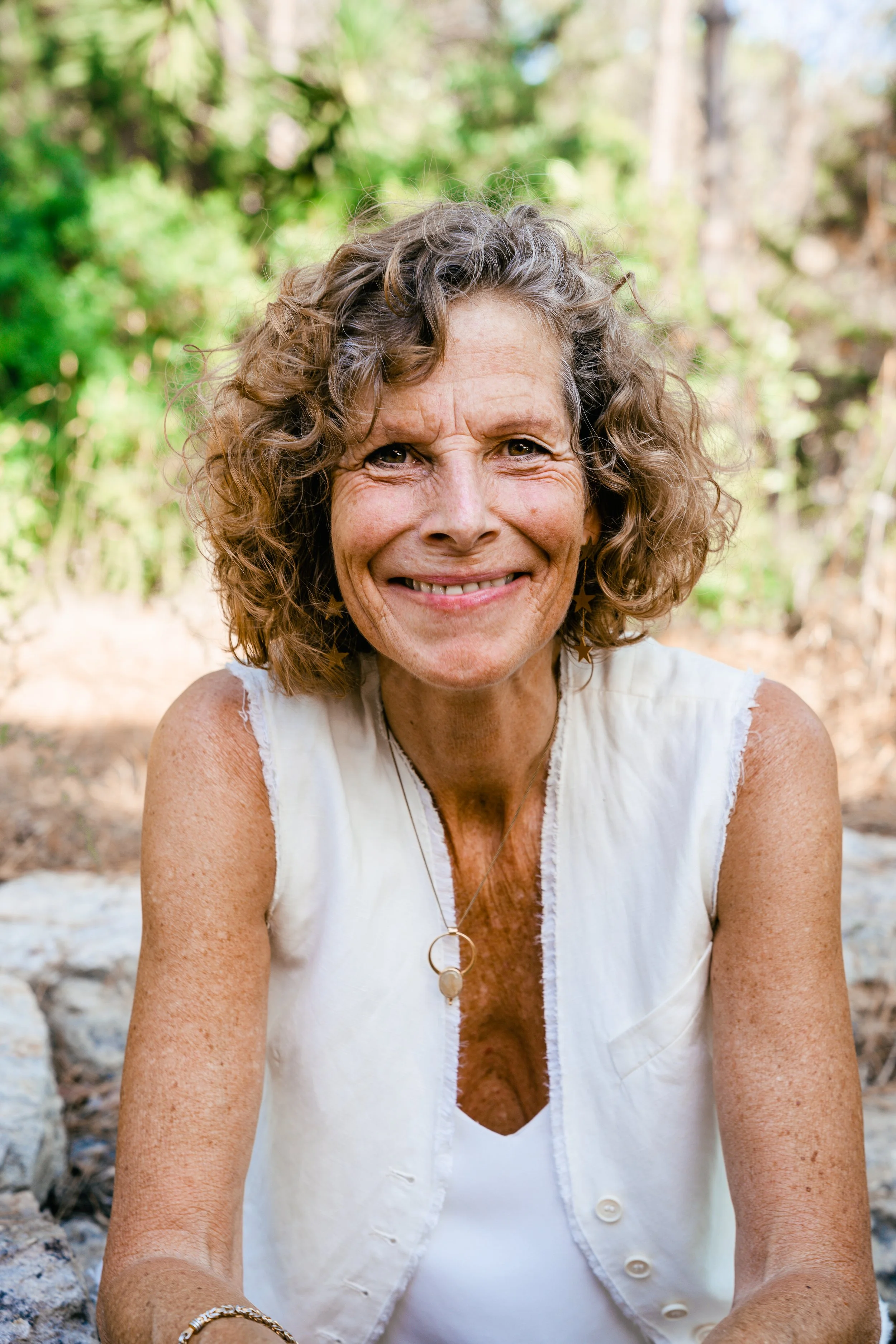 Portrait of a middle-aged woman with curly hair, smiling, outdoors with green trees in the background.