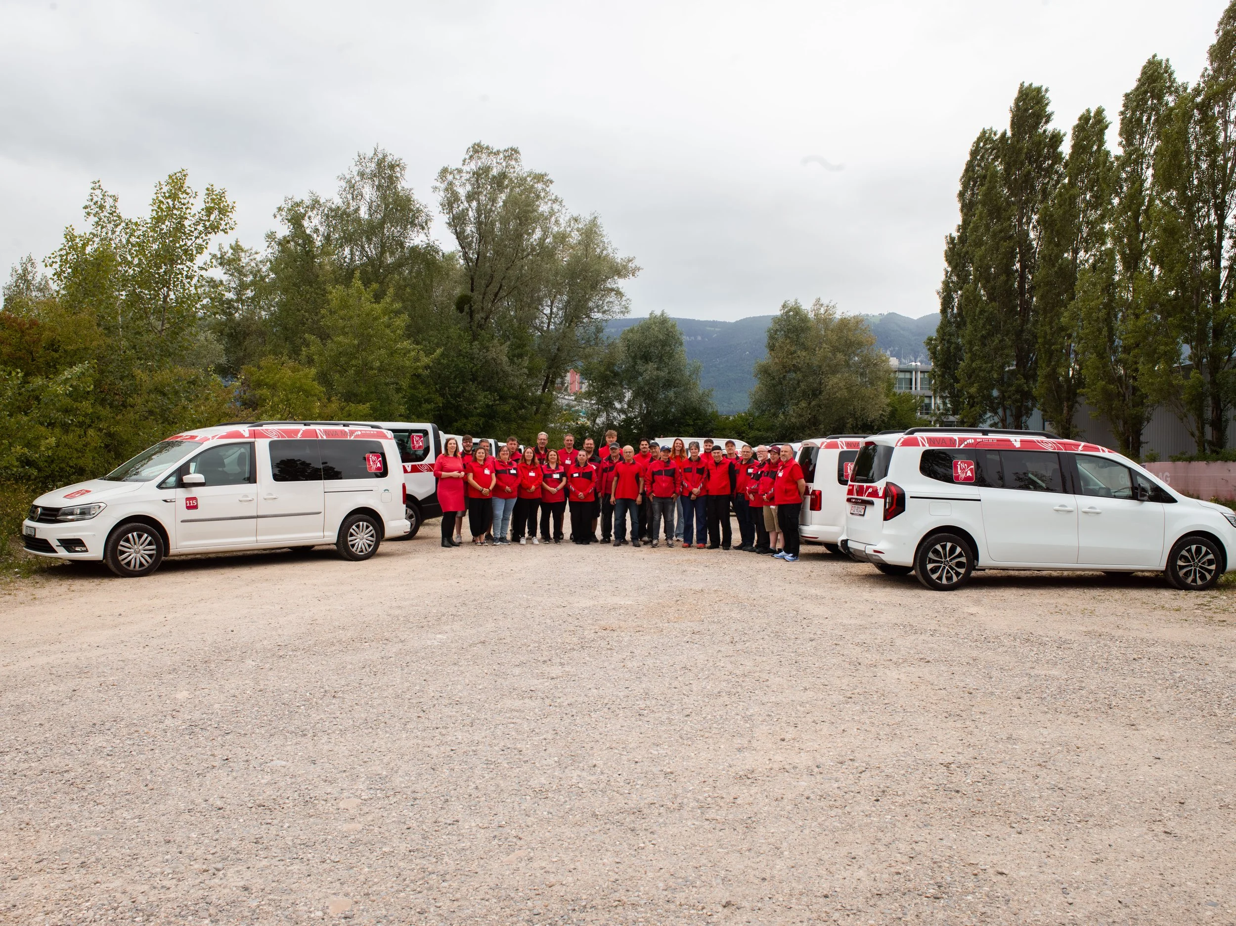 Das INVA Mobil Team, die in rot-schwarzen Jacken stehen, vor unseren Fahrzeugen stehen. Sie sind im Freien auf einer unbefestigten Fläche, umgeben von Bäumen und Bergen im Hintergrund.