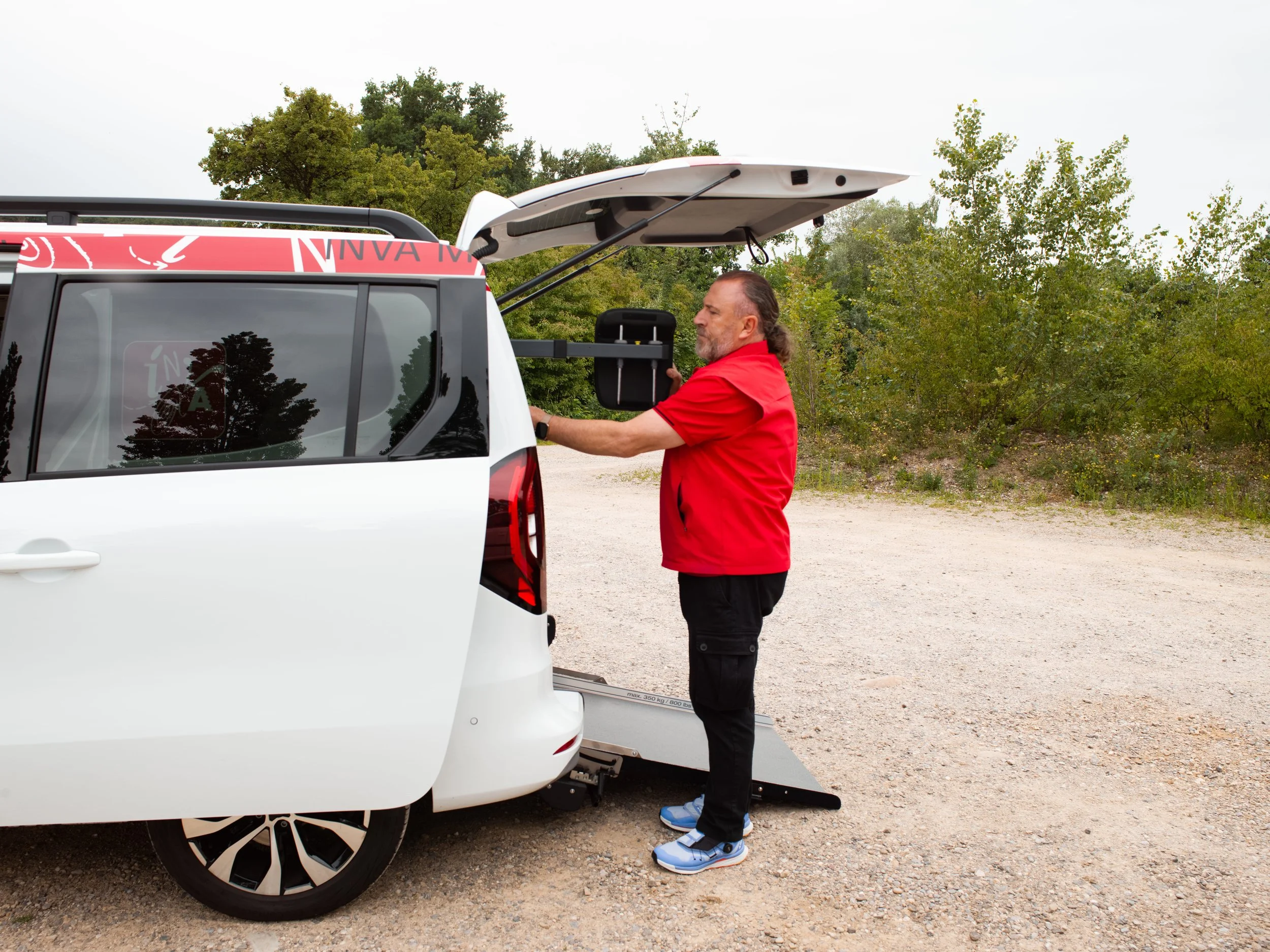 Ein Mann im INVA Mobil–Polo und schwarzen Hosen nimmt aus einem weissen Elektroauto etwas heraus.