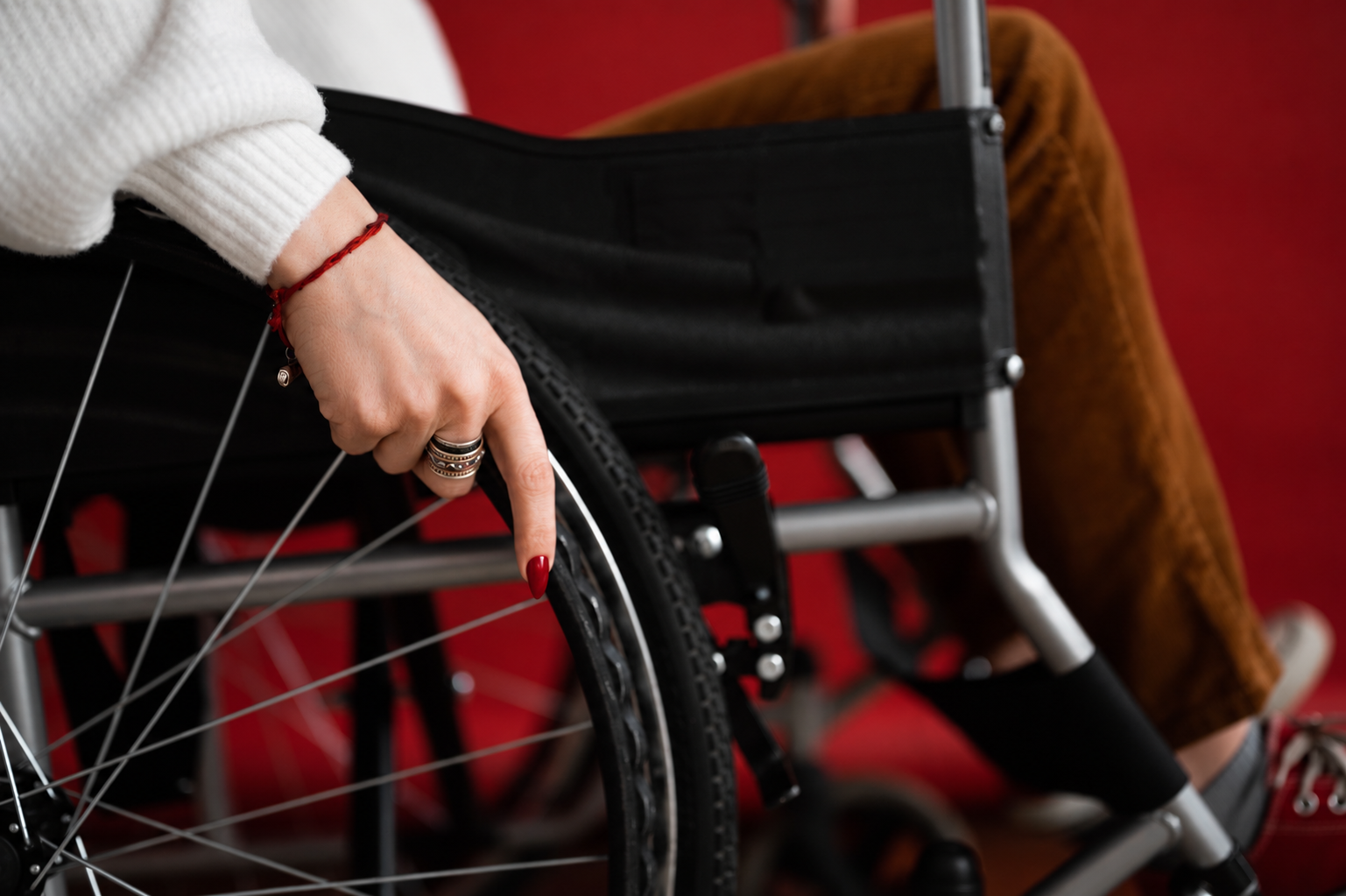 Nahaufnahme einer Hand, die an einem Rollstuhl das Rad berührt, mit rot lackierten Fingernägeln, mehreren Ringen und einem roten Armband, im Hintergrund eine Person, die im Rollstuhl sitzt, vor einem roten Hintergrund.