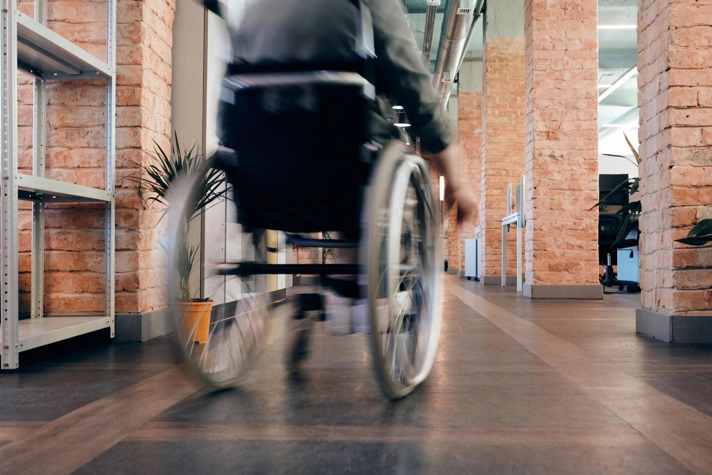 Ein Rollstuhlfahrer fährt durch eine moderne, gut beleuchtete Büroflur mit Ziegelsteinwänden und Pflanzen.