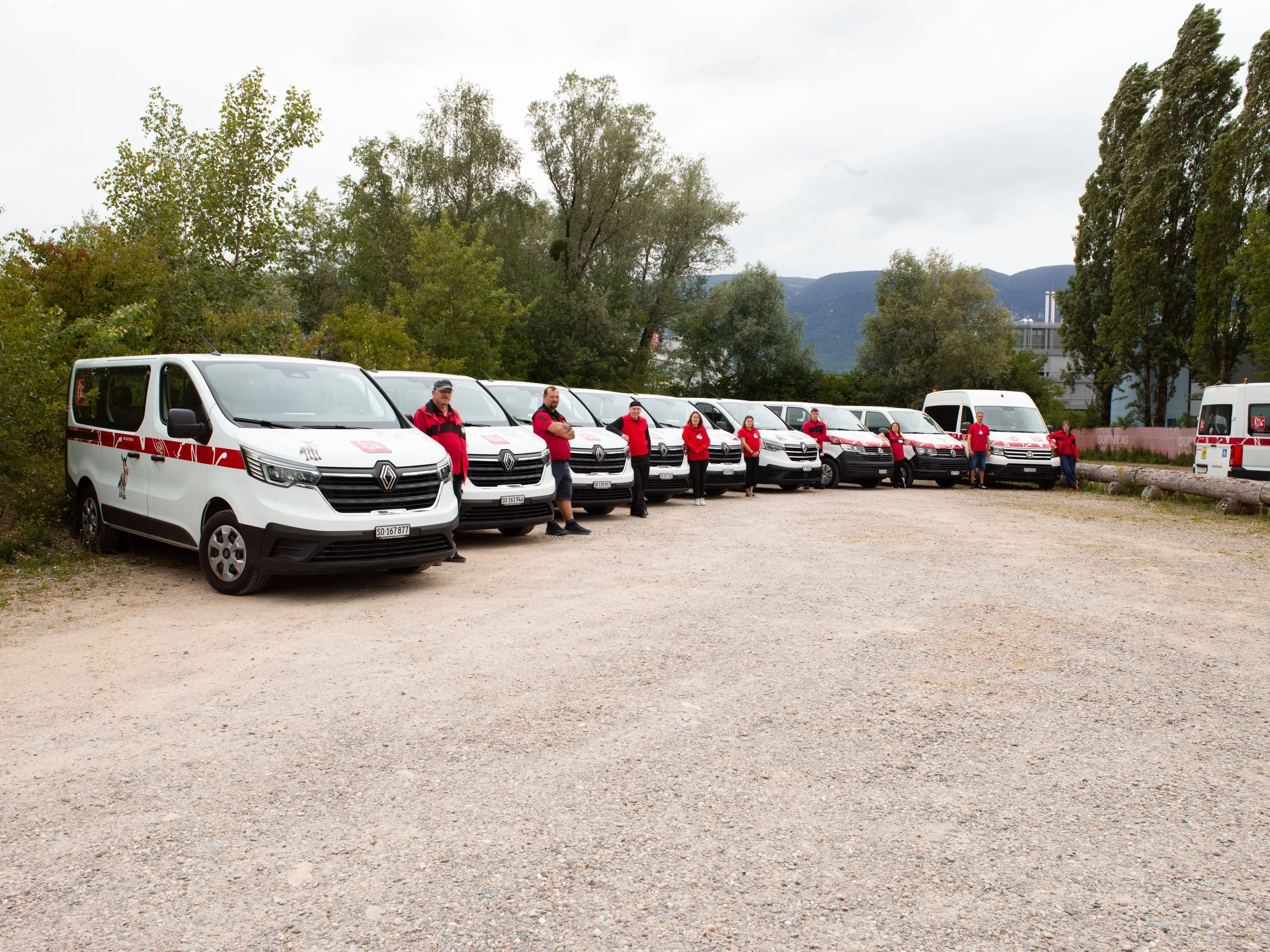 Das INVA Mobil Team steht in rote-schwarzen Jacken vor den Fahrzeugen. Im Hintergrund Bäume und Berge, bewölkter Himmel.