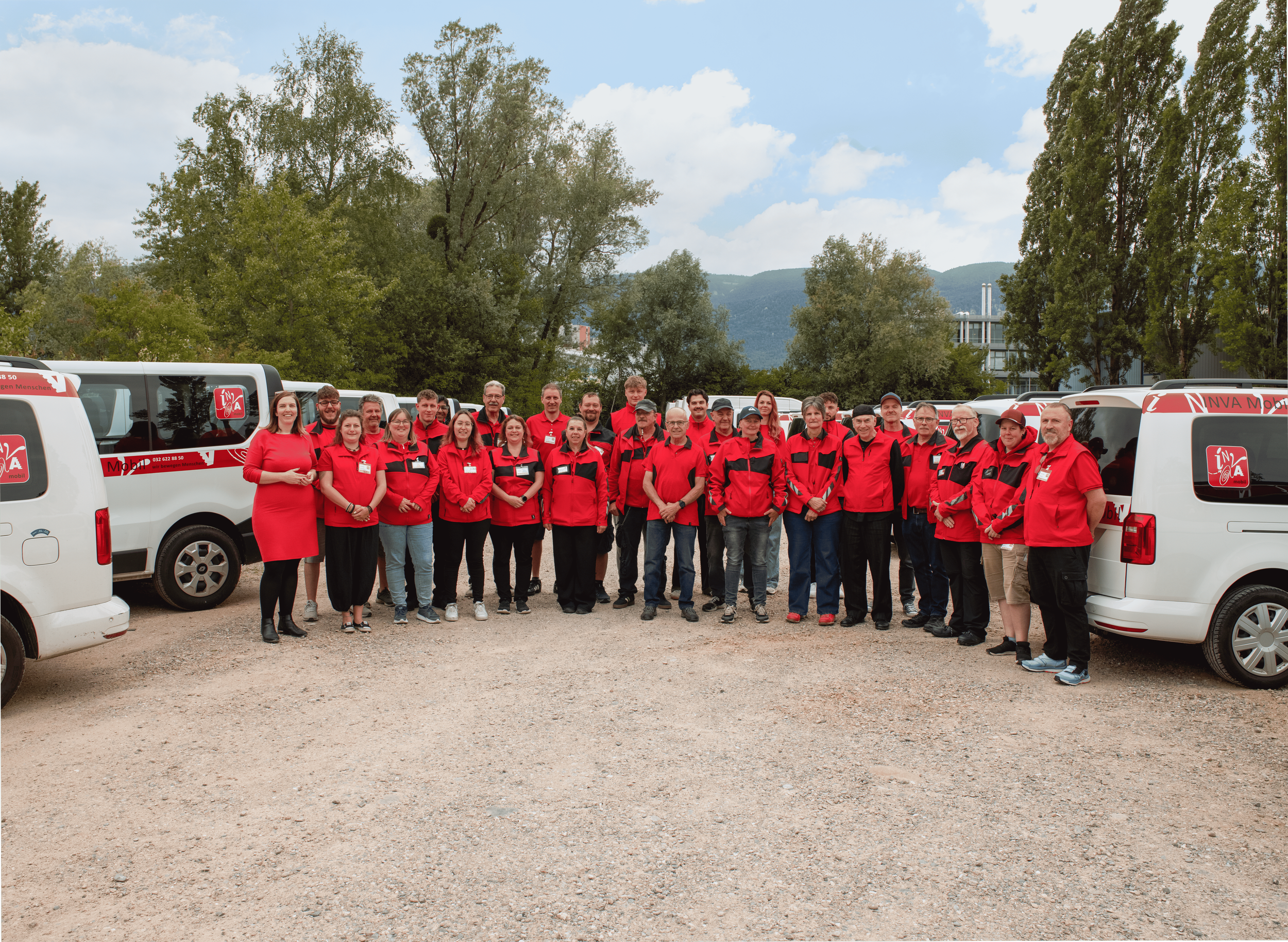 Das INVA Mobil Team steht in rote-schwarzen Jacken vor den Fahrzeugen. Im Hintergrund Bäume und Berge, bewölkter Himmel.
