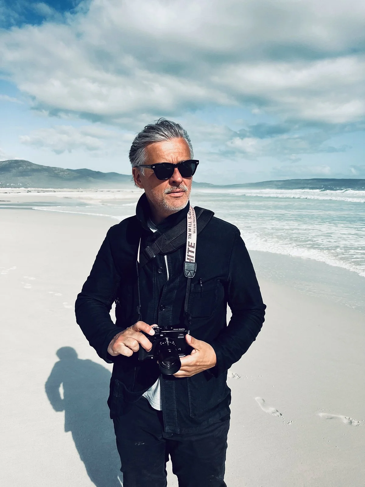Ein Mann mit grauem Haar, Sonnenbrille und dunkler Kleidung steht am Strand mit Blick aufs Meer, hält eine Kamera in der Hand.