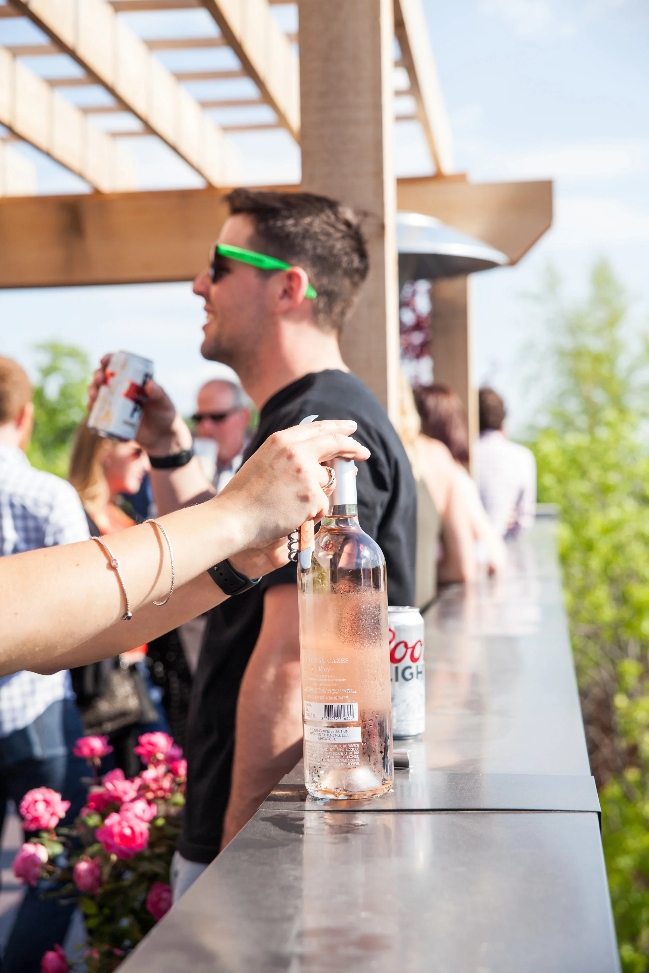 A man in sunglasses smiling with a bottle of rosé wine on a bar counter during an outdoor gathering.