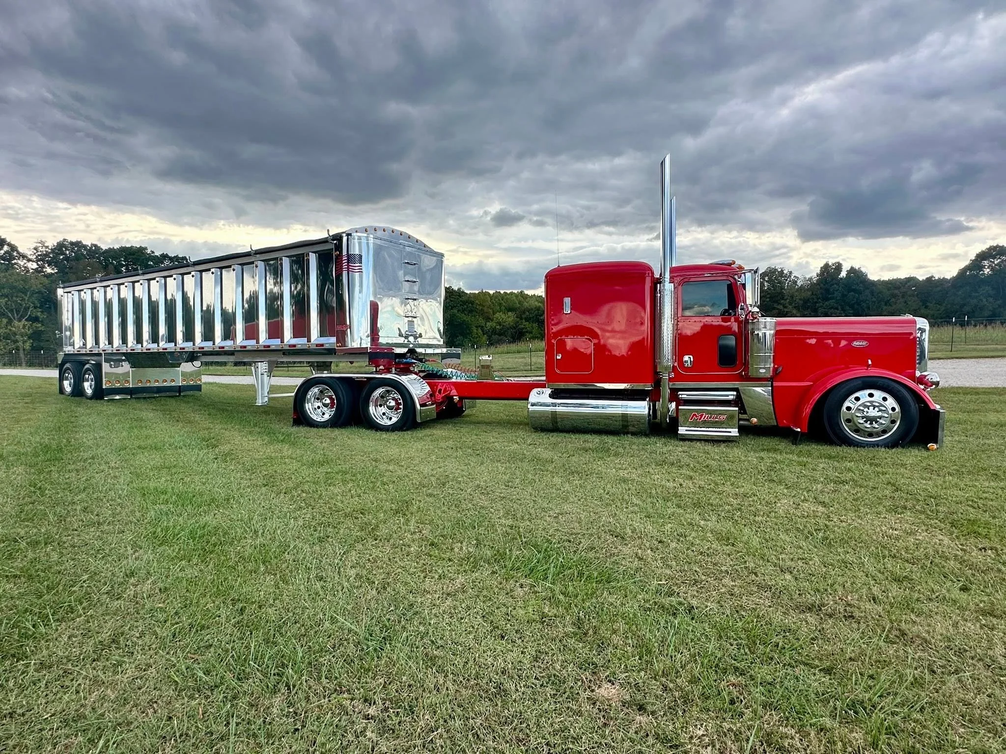 Red semi-truck with chrome accents parked on a grassy field under a cloudy sky, attached to a reflective silver trailer.