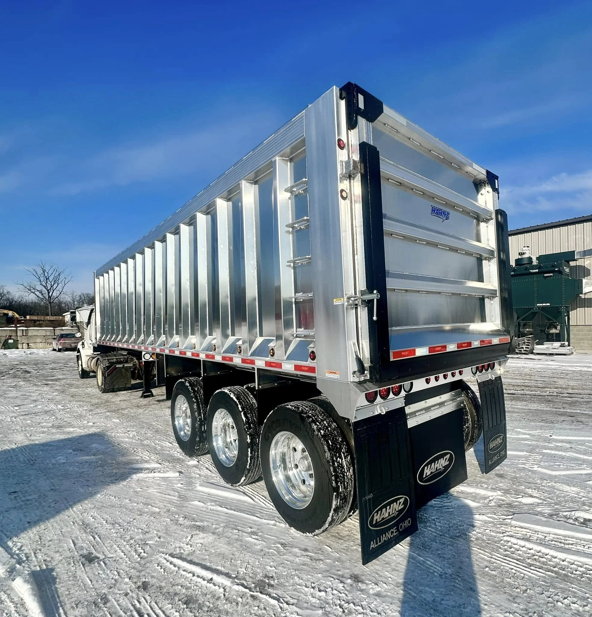 Silver aluminum flatbed trailer parked on snowy ground with a building in the background, clear blue sky, and a leafless tree.