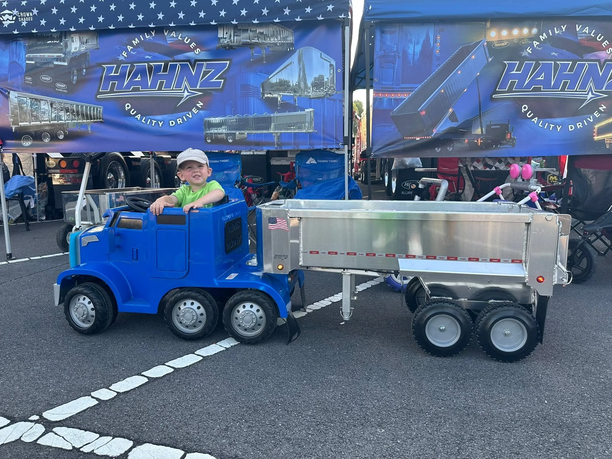 A young boy sitting in a small blue toy truck with a striped black and grey handlebar, surrounded by trailers and tents, at an outdoor event.