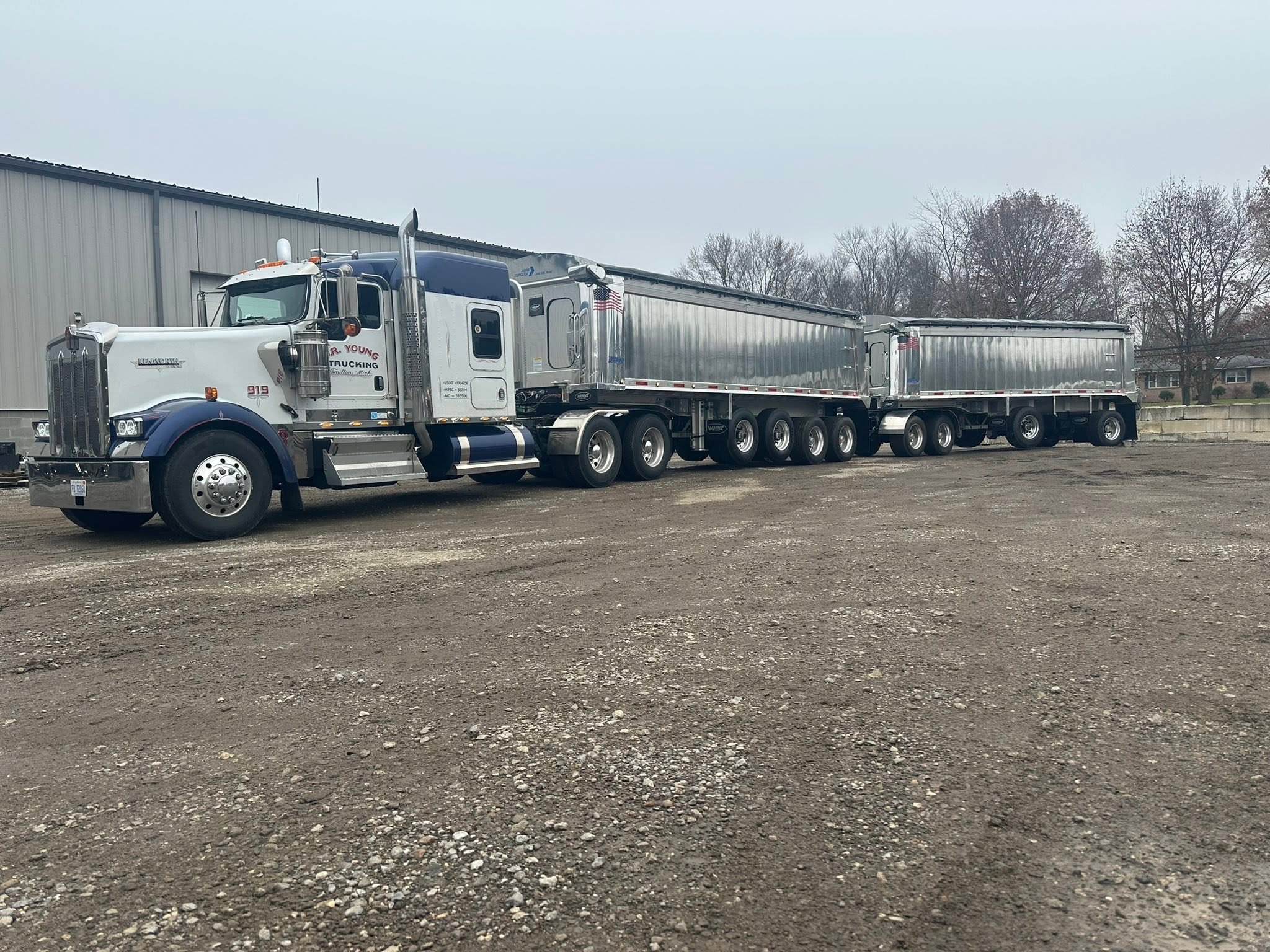 A large semi-truck with a blue and white cab and silver trailers parked outdoors on a gravel surface, with trees and a building in the background.