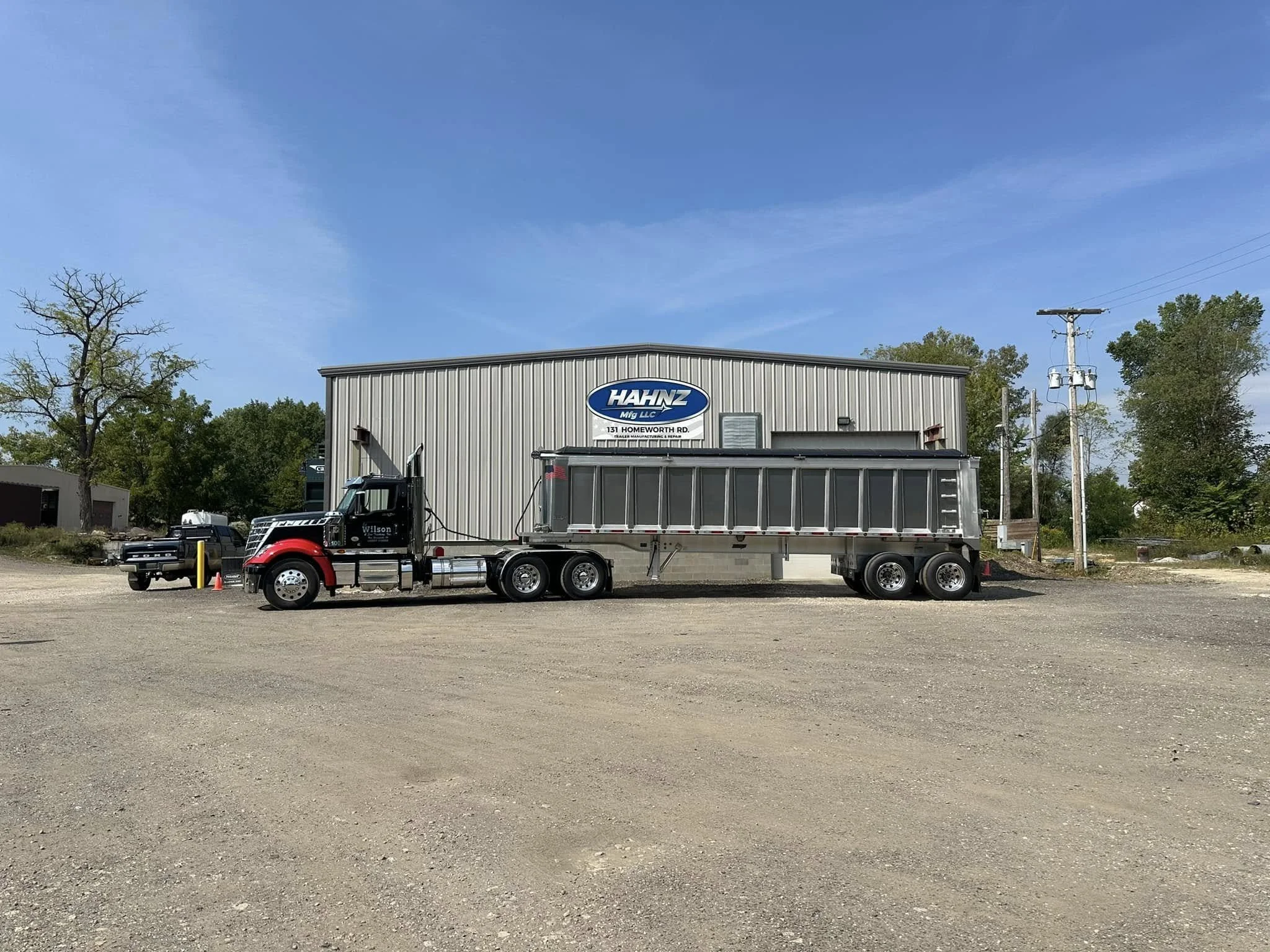 A large industrial building with a sign that reads "HAHNZ Mfg LLC" located at 131 Homeworth Road. There is a black semi-truck with a flatbed trailer parked in front of the building. The sky is mostly clear with some wispy clouds, and there are trees and power lines in the background.