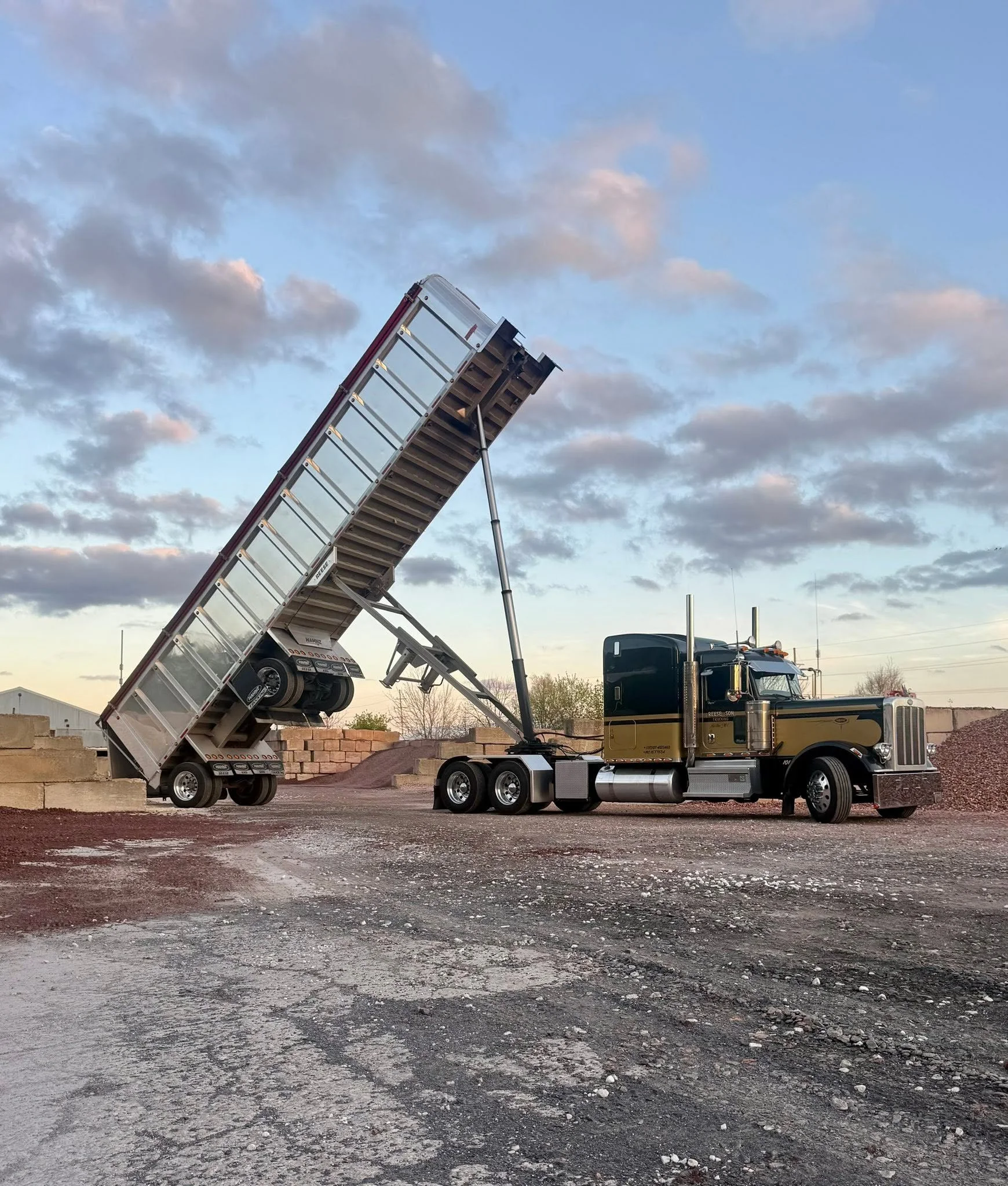 A large black semi-truck with a shiny gold stripe, with an extended ladder on the back of its trailer, on a gravel lot with brick walls and a partly cloudy sky in the background.
