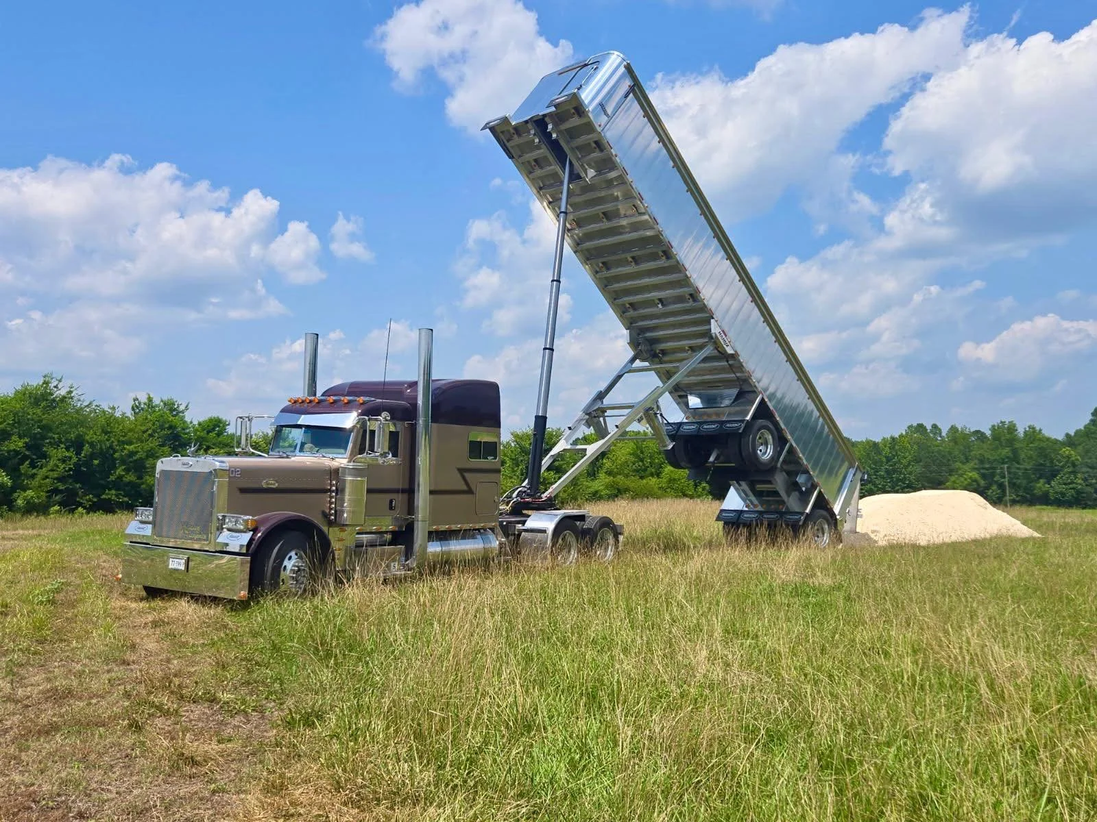 A large semi-truck with a reflective metallic trailer is positioned in a grassy field. The trailer is raised at an angle, and a small car is visible underneath it. The scene is outdoors on a bright, partly cloudy day with trees in the background.