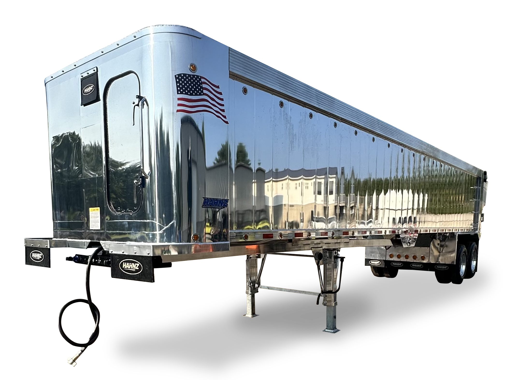 A shiny silver enclosed trailer with an American flag decal on the side. The trailer has a black door handle, a small window, and additional branding and safety labels. It is supported by metal landing gear and connected to a hitch, with tires visible underneath.