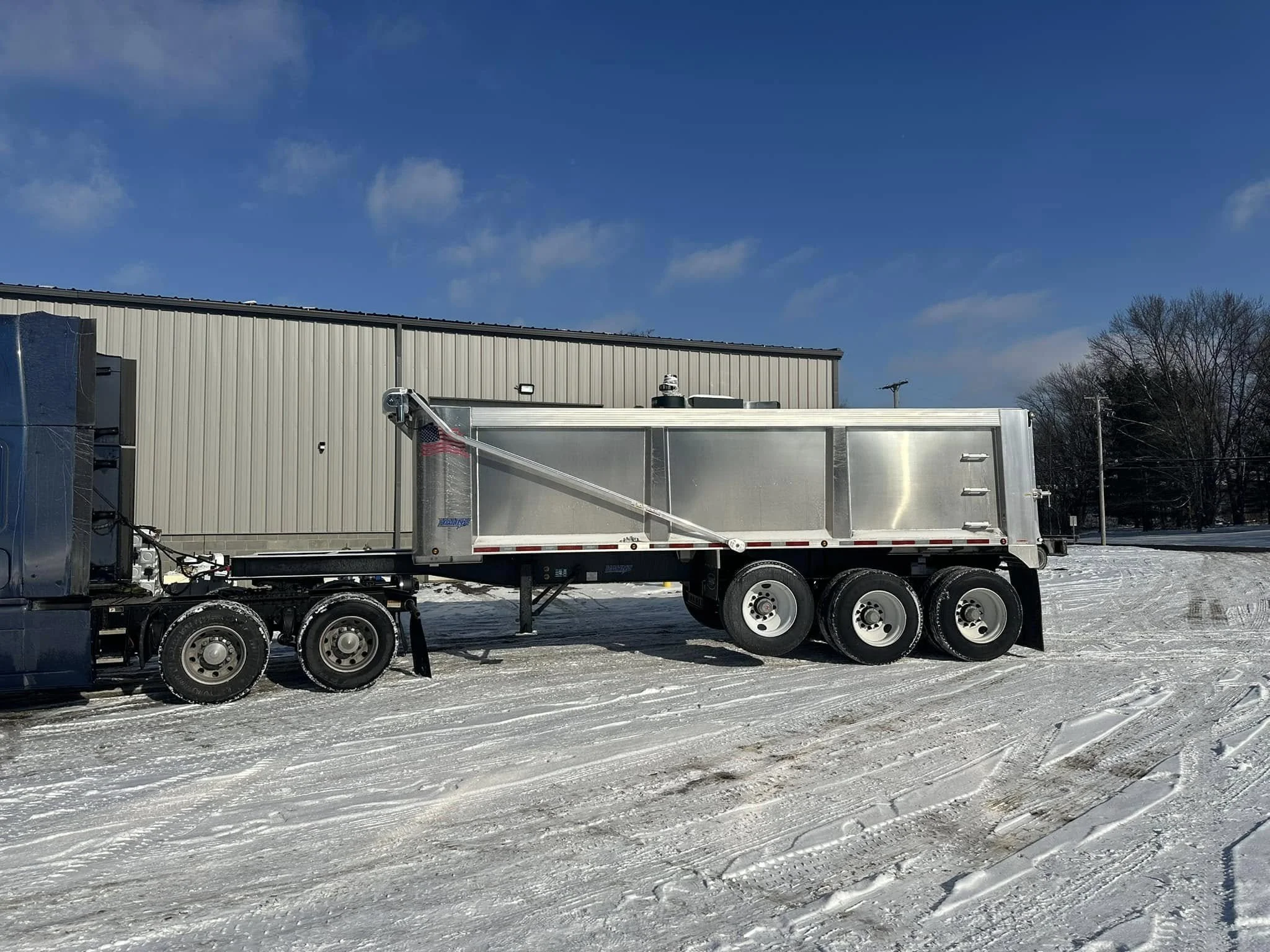 A large white truck with flatbed trailer parked on snow-covered ground outside a beige industrial building under a blue sky with scattered clouds.