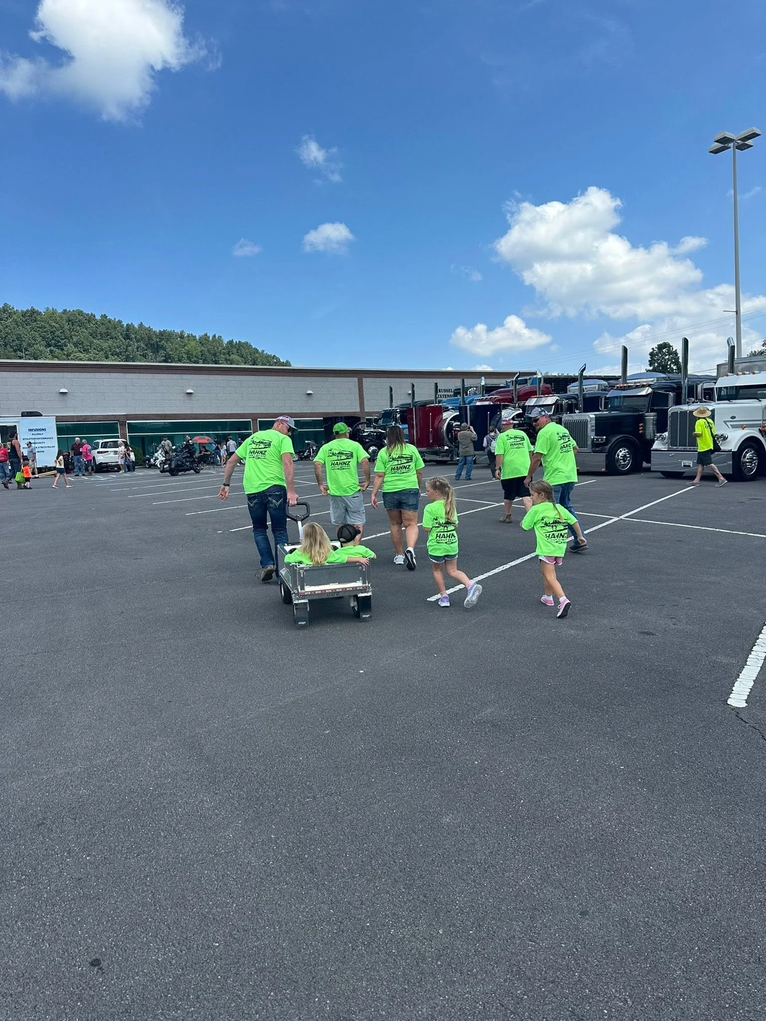 People wearing bright green shirts walking in a parking lot near a row of semi trucks, with a building, a tree-covered hill, and a bright blue sky with scattered clouds in the background.