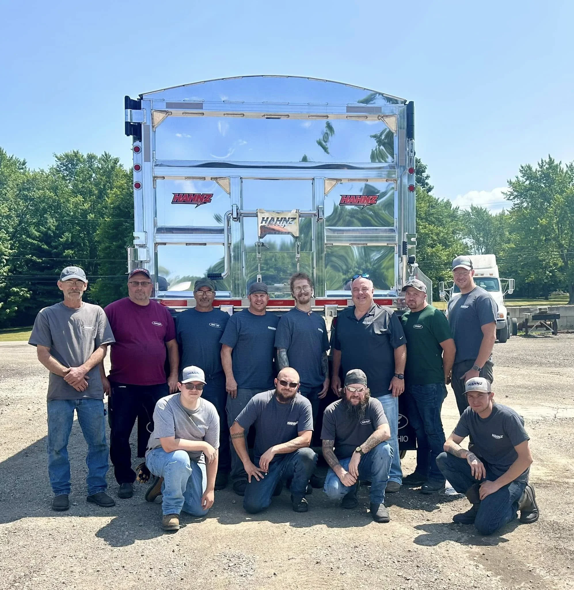 A group of men standing outdoors in front of a large, reflective trailer. The men are smiling and wearing casual clothing, some with hats and sunglasses. The background includes green trees and a clear blue sky.