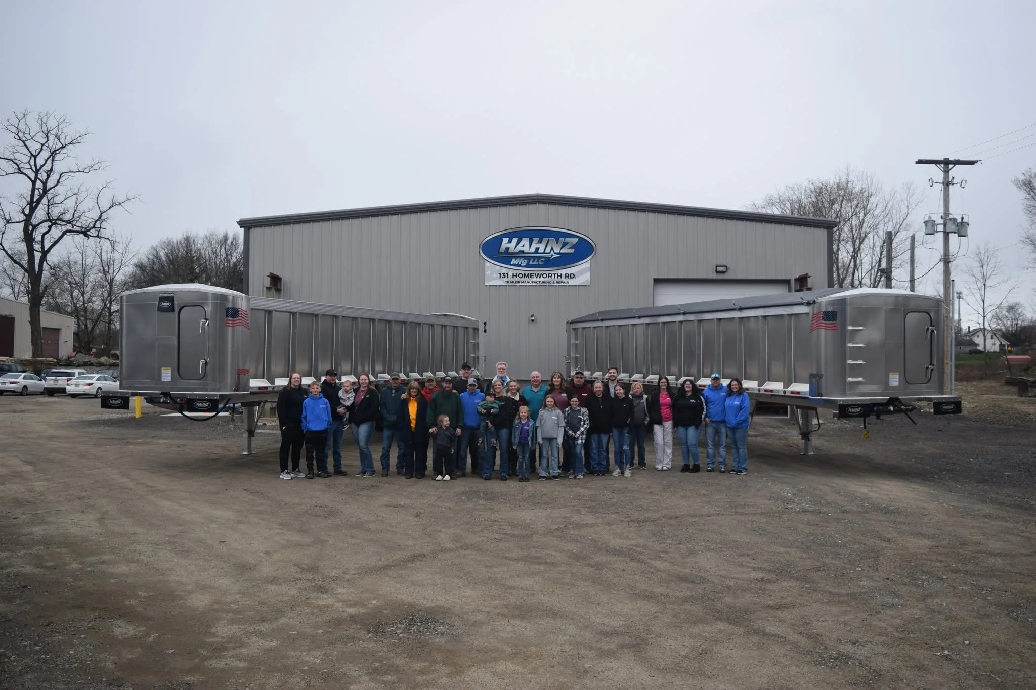 A group of people standing in front of two large trailers outside a building with a sign that reads 'Hahnz Mig LLC, 131 Homeworth Rd, Trailer Manufacturing & Repair.' The sky is overcast, and there are trees and cars in the background.