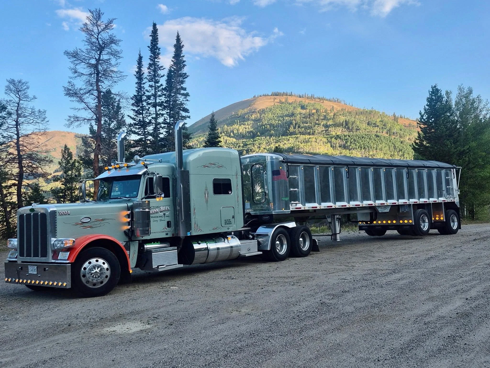 A silver semi-truck with a black trailer and orange accents parked on a gravel lot, with a backdrop of green, forested hills and tall trees under a blue sky with some clouds.