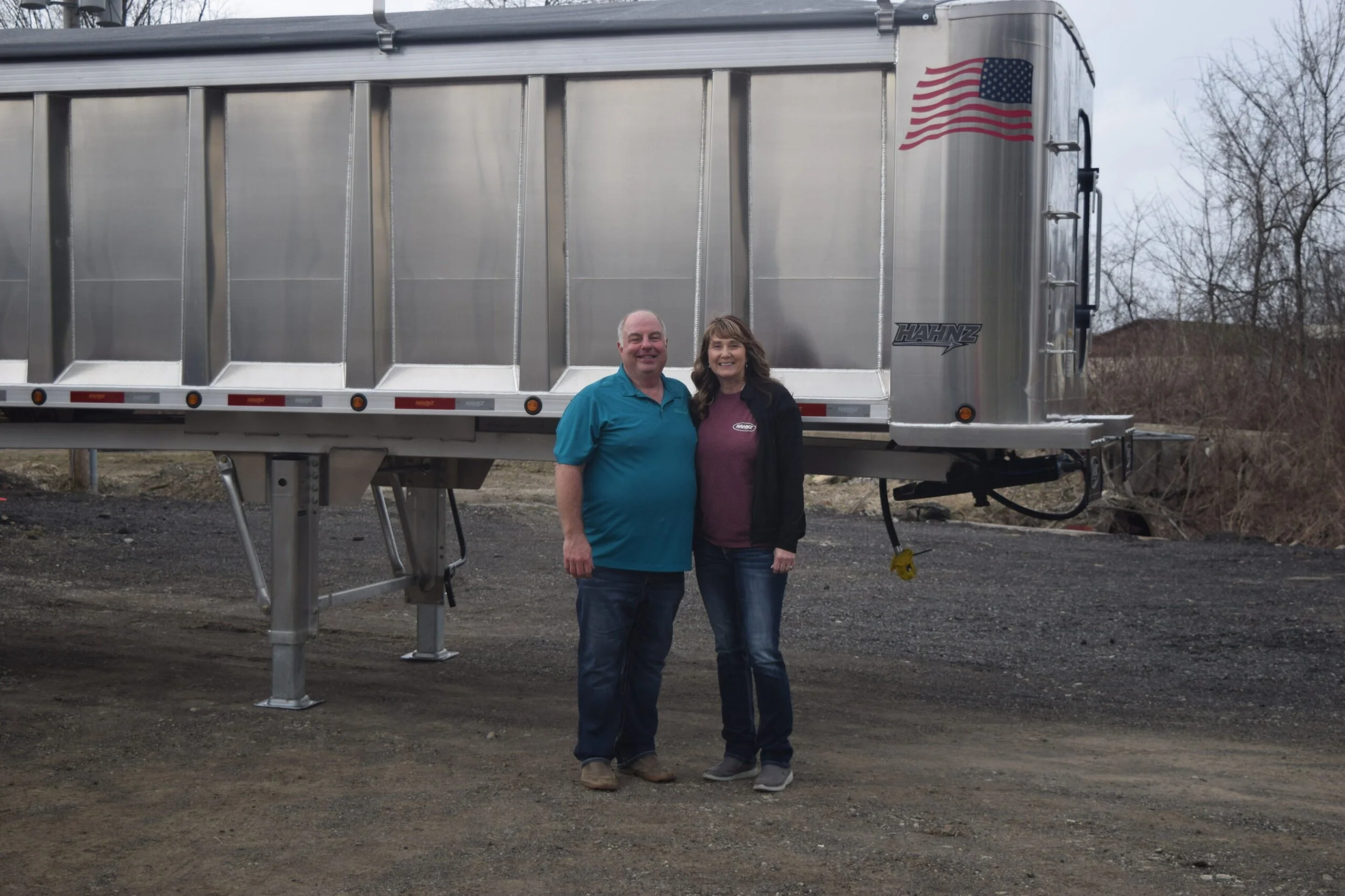 Two people, a man and a woman, smiling and standing close together in front of a large silver trailer with an American flag decal on the upper right corner. The background shows leafless trees on a cloudy day.