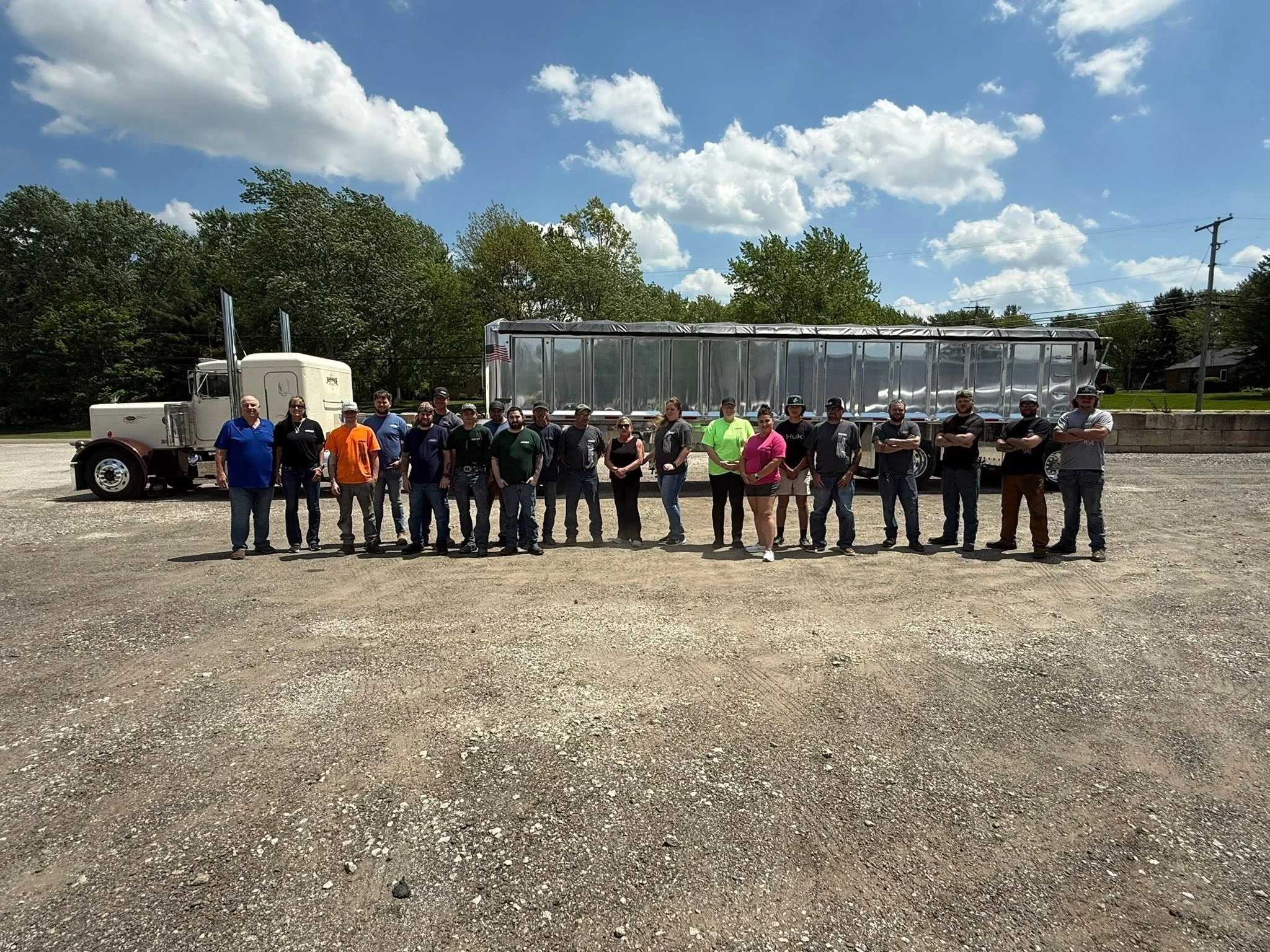 Group of people standing outside in front of a large truck with a reflection on its surface, under a sky with scattered clouds and trees in the background.