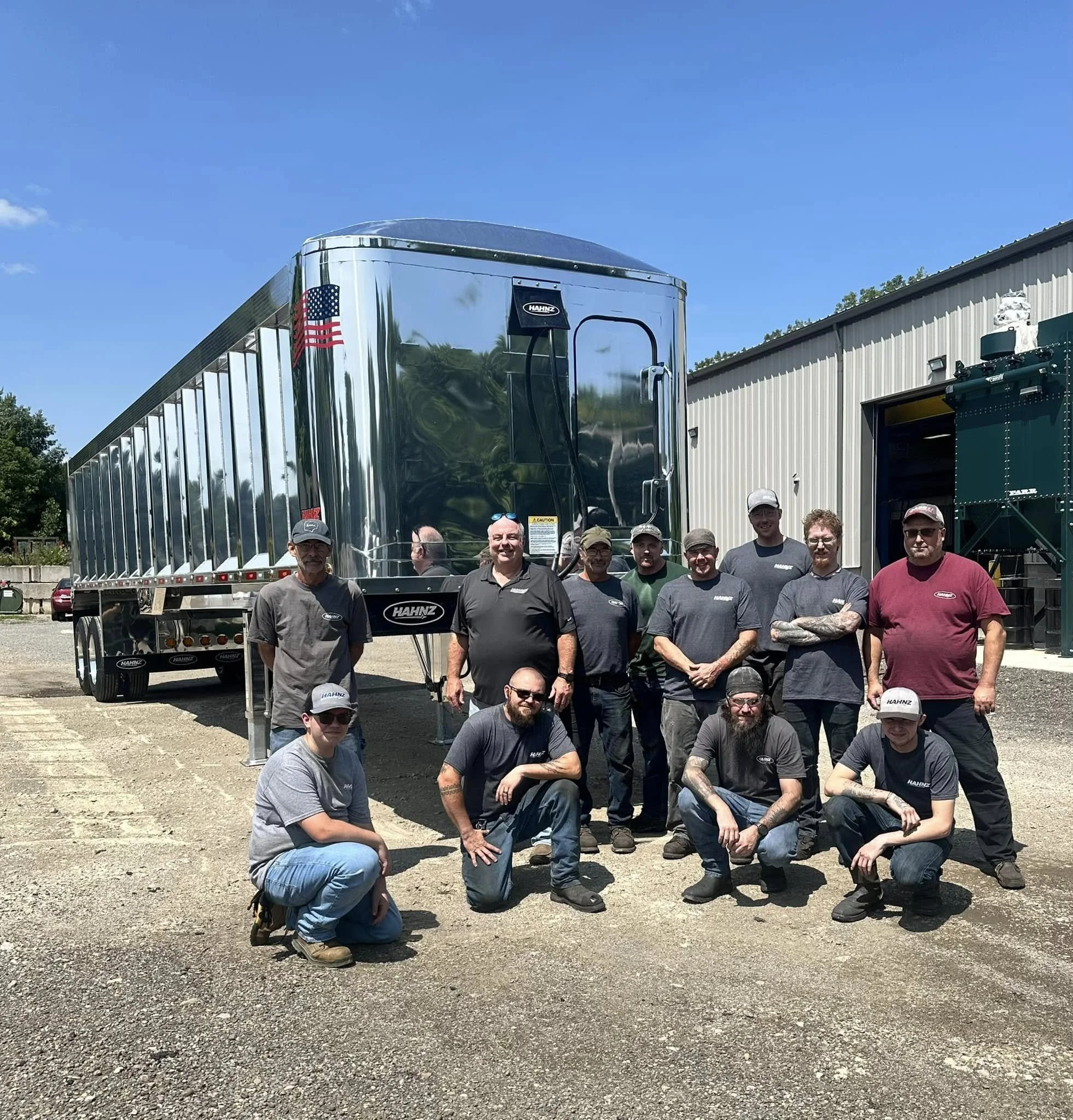 Group of men standing and crouching outdoors in front of a large shiny metallic trailer with Kaiser logo, some are wearing hats with HawHz branding, under a clear blue sky.