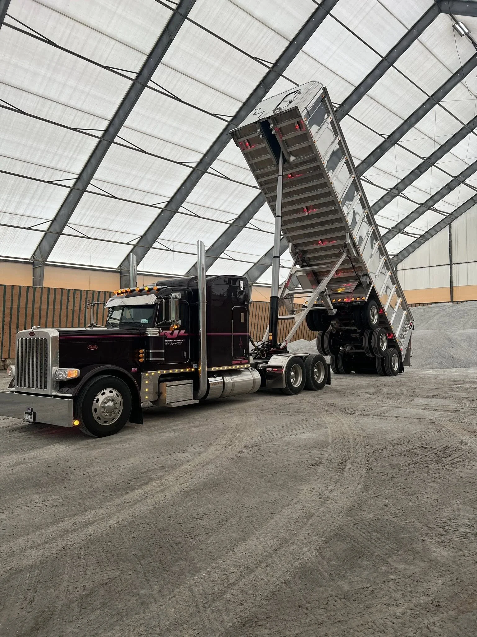 A semi-truck with a black cab and a silver flatbed trailer is inside a large industrial building with a translucent curved roof. The trailer's ramp is down and inclined, and the ground is covered in dirt or gravel.