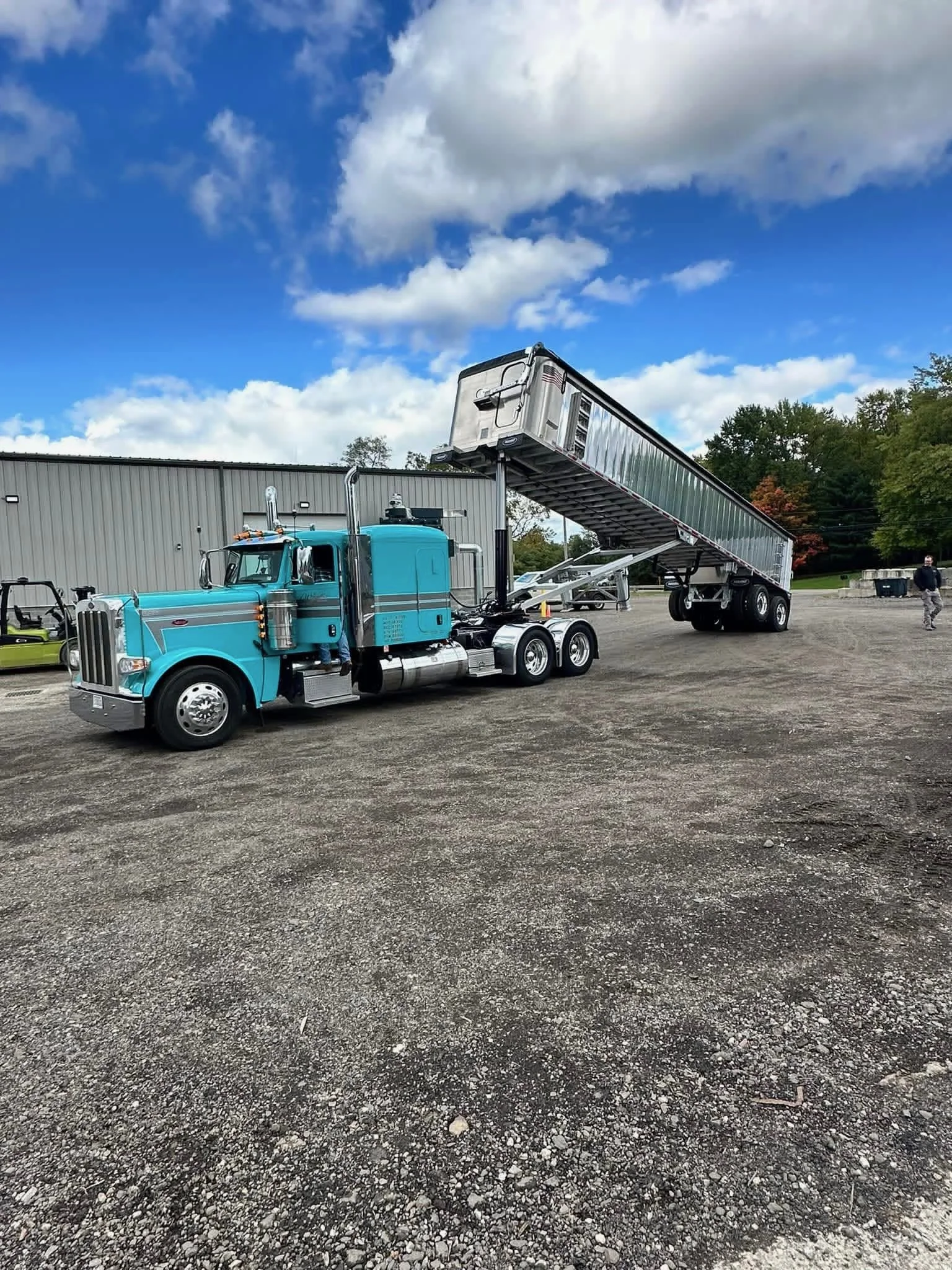 A blue semi-truck with a raised trailer parked on a gravel lot outside a large building, with a person standing nearby and trees in the background.
