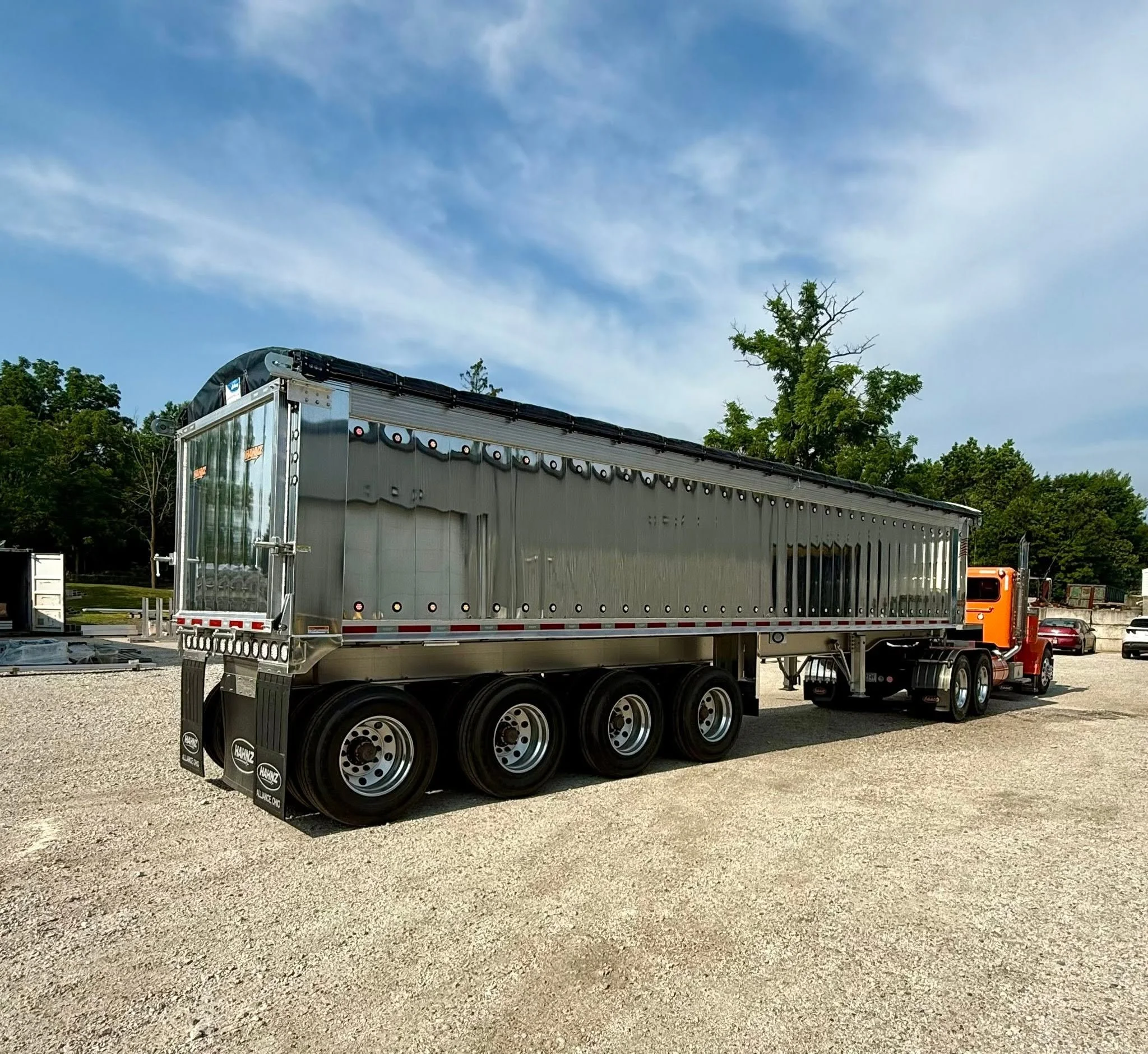 A large semi-trailer truck parked outdoors on a gravel lot with trees and a blue sky with some clouds in the background.