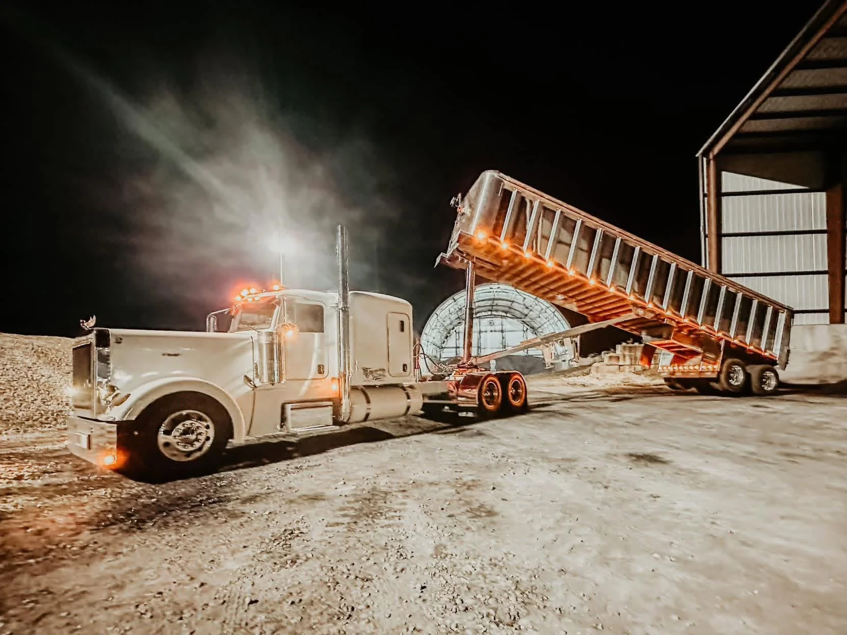 Nighttime construction site with a white semi-truck and a trailer with an inclined ramp, orange safety lights, and a large arched structure in the background under a dark sky.