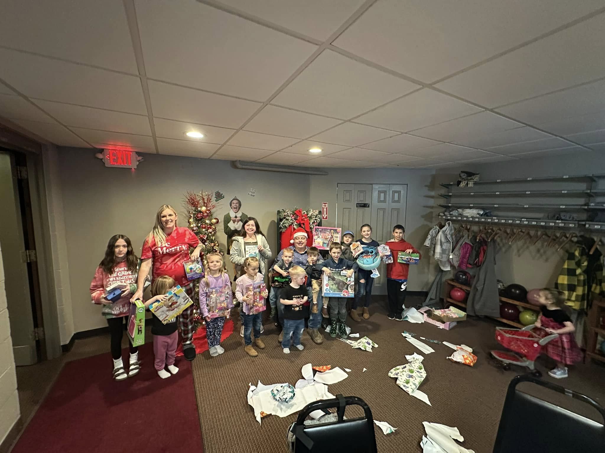 Children and adults gathered in a room celebrating Christmas, with torn wrapping paper and gifts scattered on the floor, a decorated Christmas tree, and a festive atmosphere.