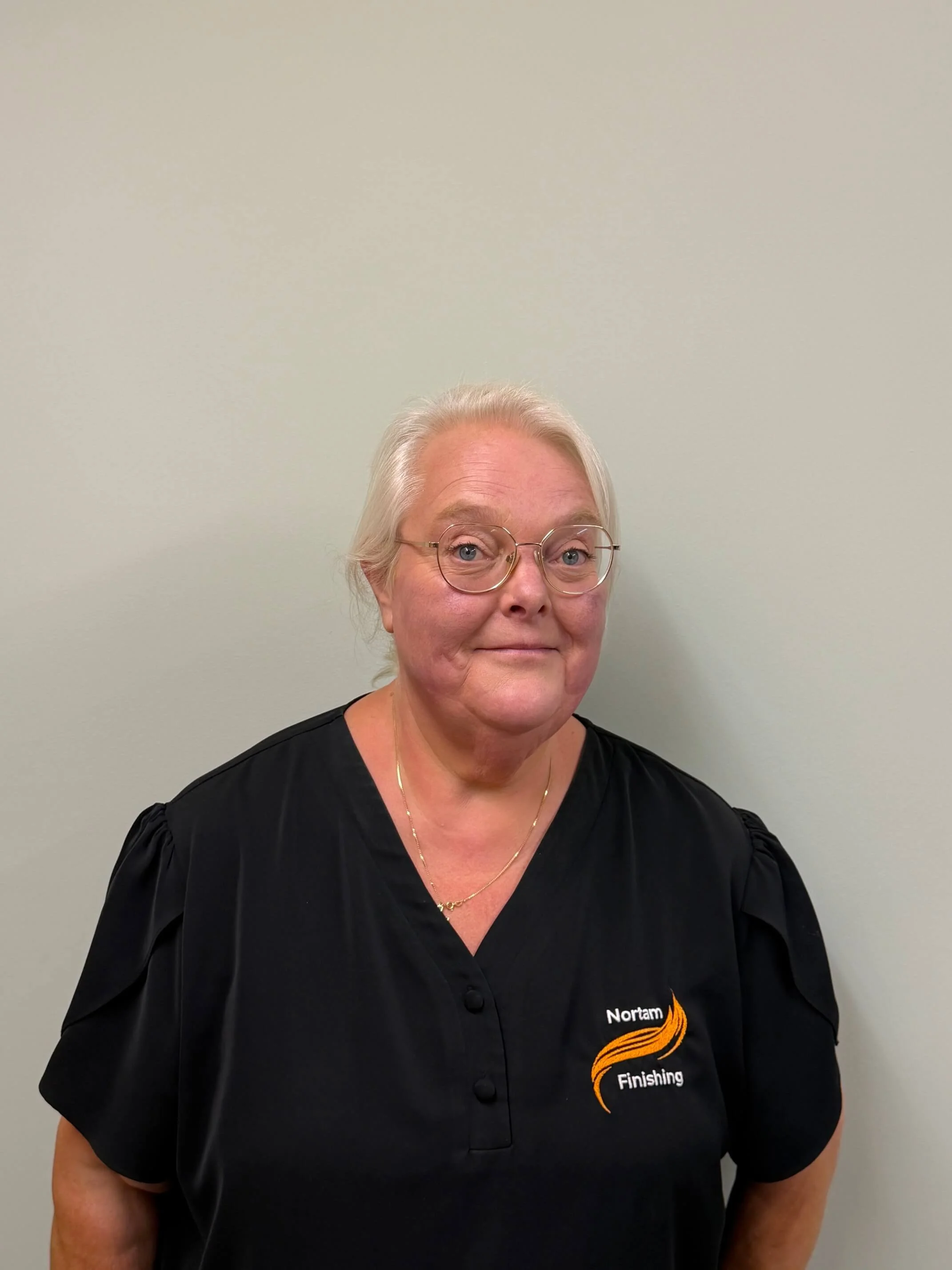 A middle-aged woman with blonde hair and glasses standing against a plain wall, wearing a black uniform shirt with a logo that reads "Nortam Finishing".