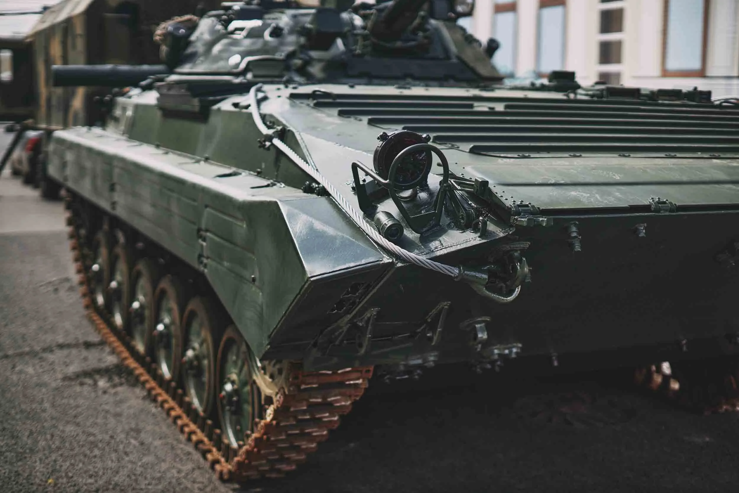 Close-up of a military tank's front section with green armor, tracks, and various equipment on top, parked on an urban street with buildings in the background.