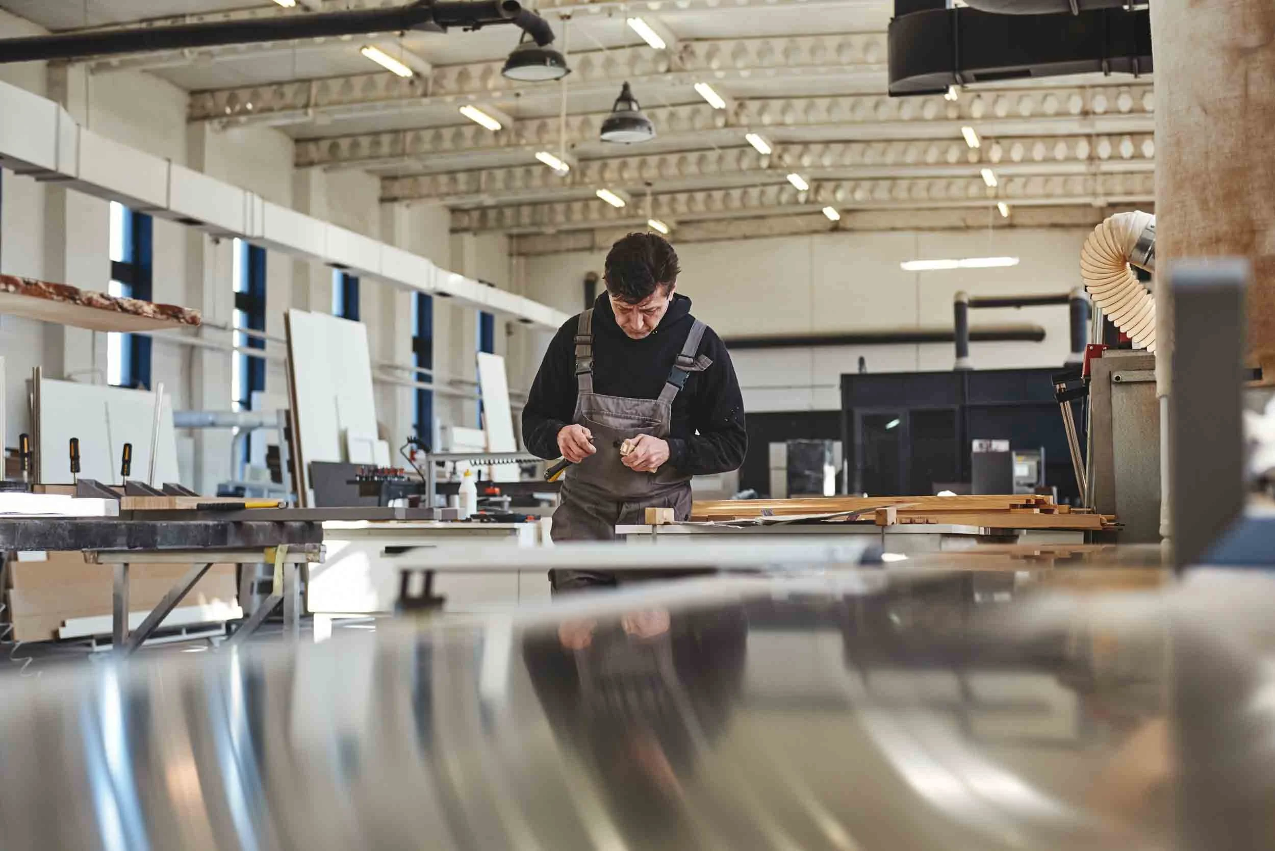 A man wearing a black hoodie and gray apron working on a woodworking project in a spacious workshop with wood beams and large windows.
