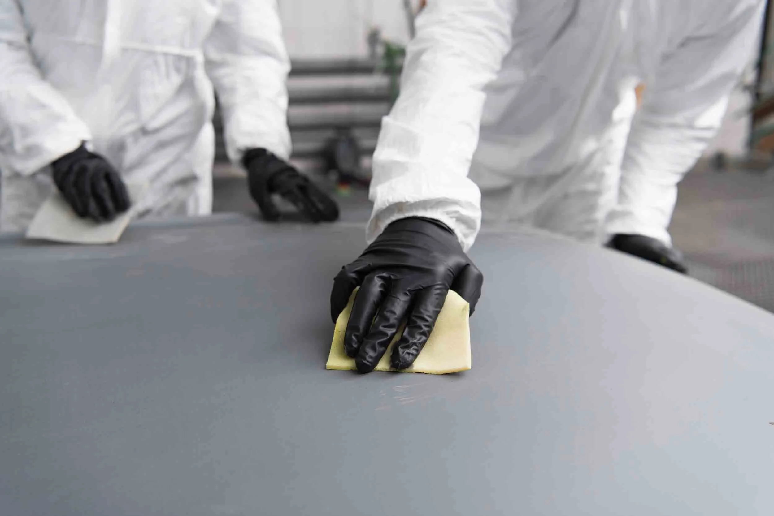 People wearing white protective suits and black gloves cleaning a large gray surface with yellow sponges.