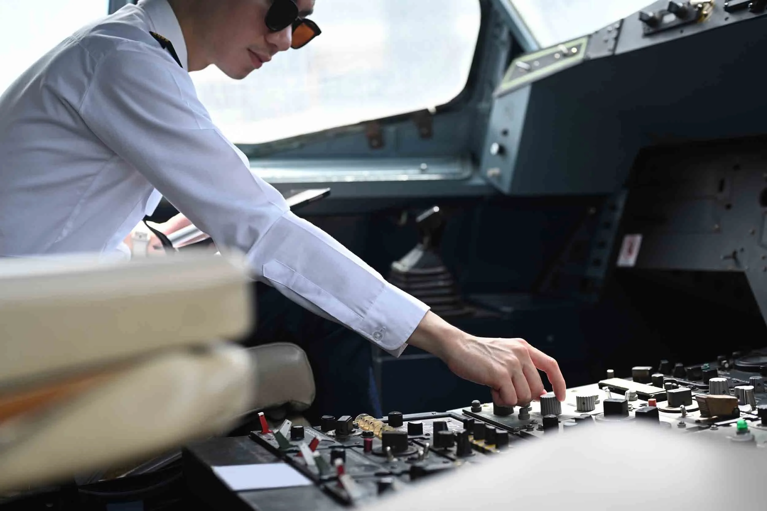 A person wearing a white shirt and sunglasses operating sound or music mixing equipment with various knobs and sliders inside an aircraft cockpit.