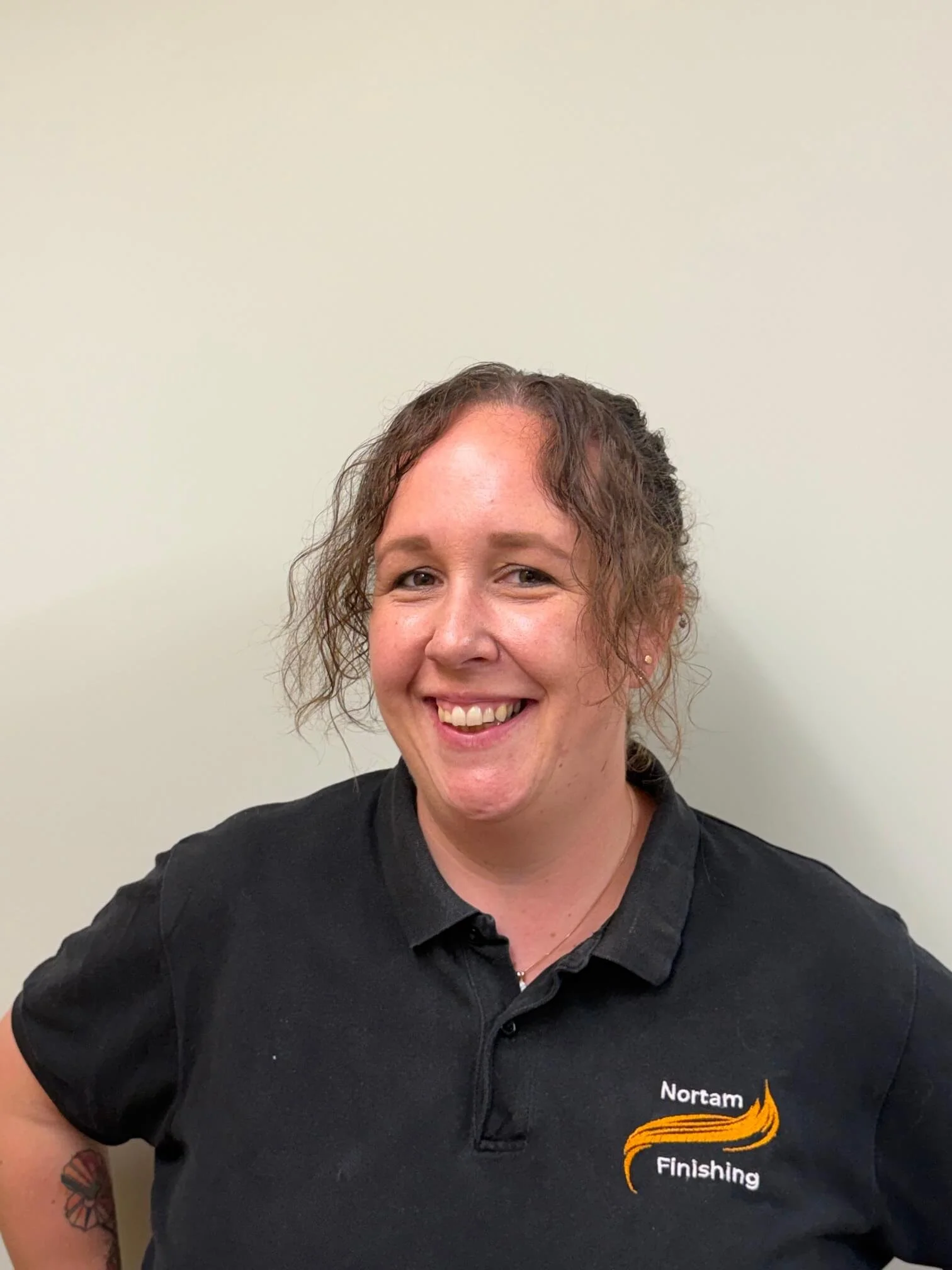 A woman with curly hair wearing a black polo shirt smiling, against a plain light-colored wall.