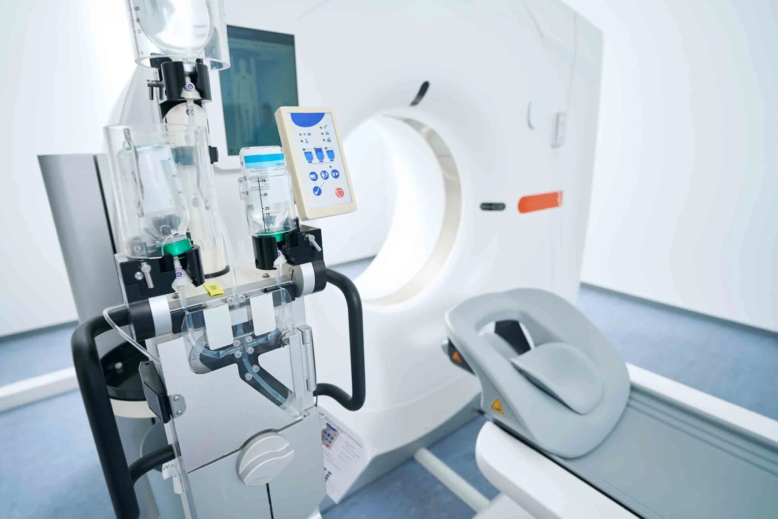 A medical imaging room featuring a large MRI scanner with a white exterior, an open circular opening, and a control panel nearby. There are various medical devices and containers with liquids placed on a stand adjacent to the MRI machine.