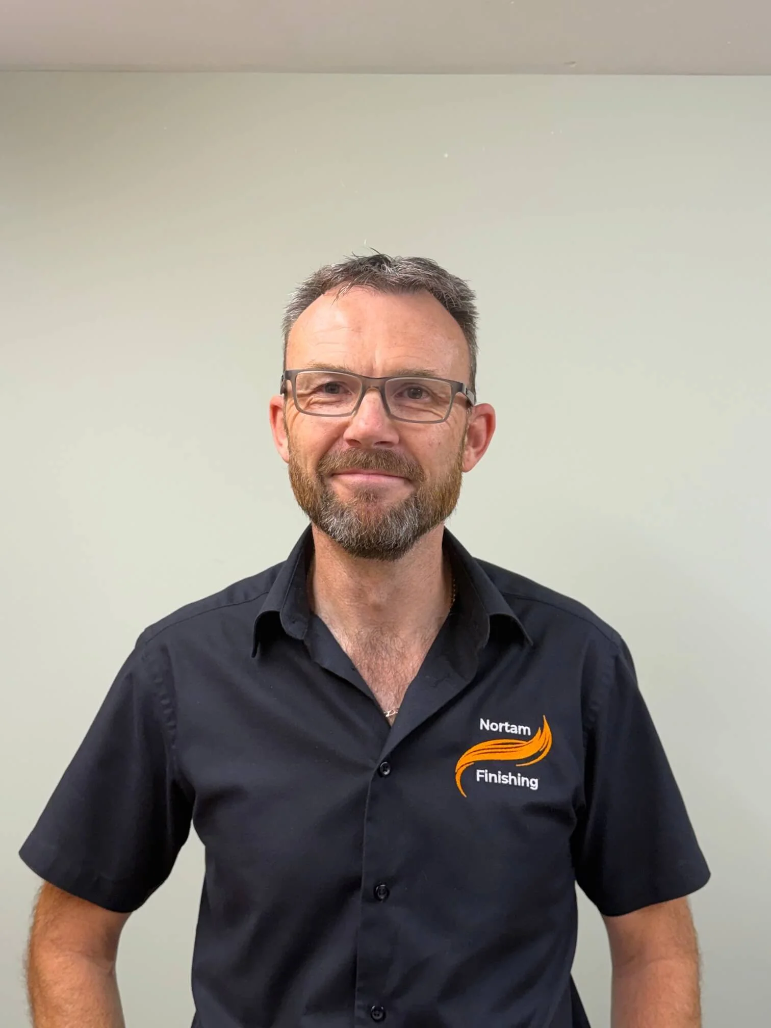 A man with short hair, glasses, and a beard wearing a black shirt with the logo 'Nortam Finishing' standing against a plain light-colored wall.