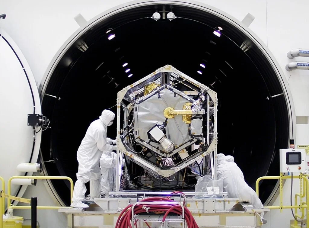 Scientists in protective suits working on a satellite inside a large cleanroom or testing chamber.