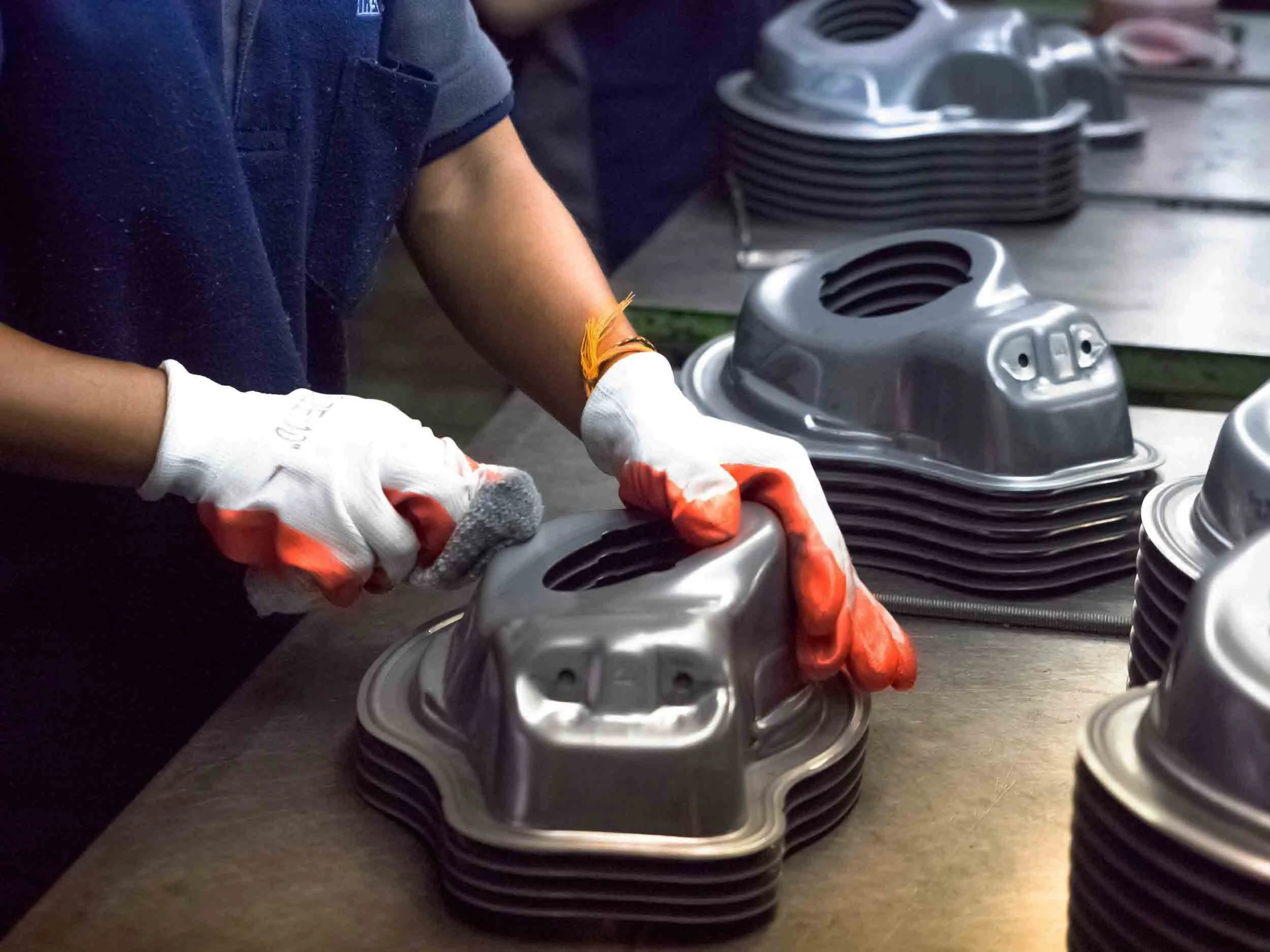 Close-up of a person wearing gloves working with engine parts on a worktable, with several silver machine components stacked in the background.