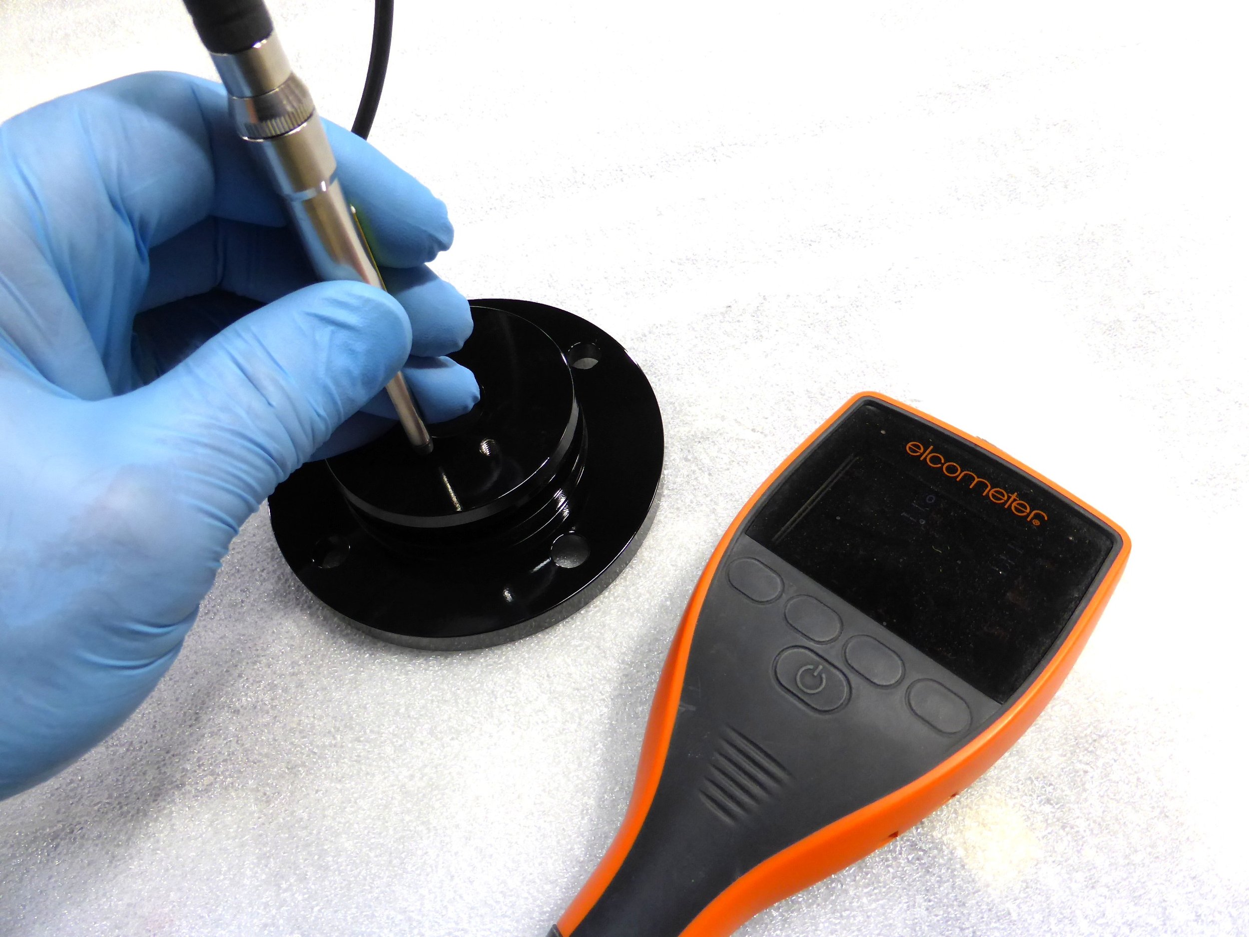 A person wearing blue gloves holding a small metallic object over a black circular base. There is an orange electronic device labeled 'elcometer' and a digital meter on the right side of the image.